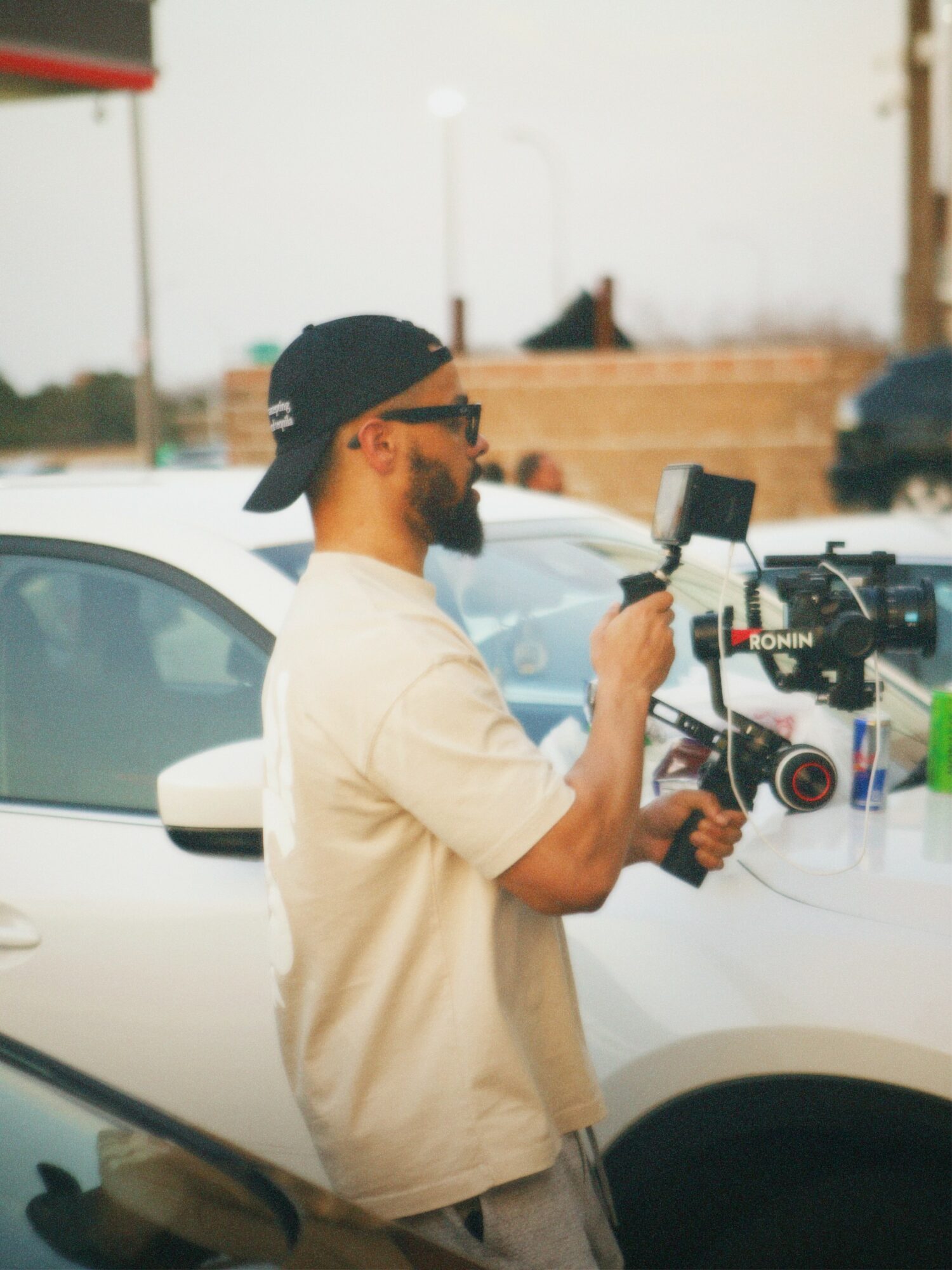 Man with a beard, wearing a black cap backwards and sunglasses, holding a camera on a white car, outdoors.
