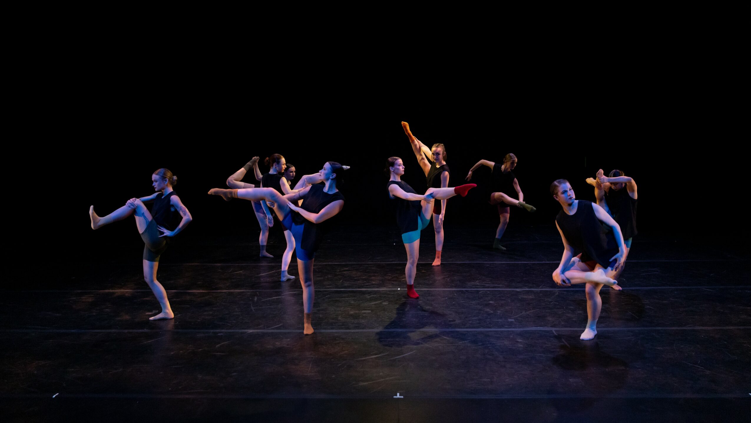 Group of dancers performing on stage with black background, some balancing on one leg, others in various poses.