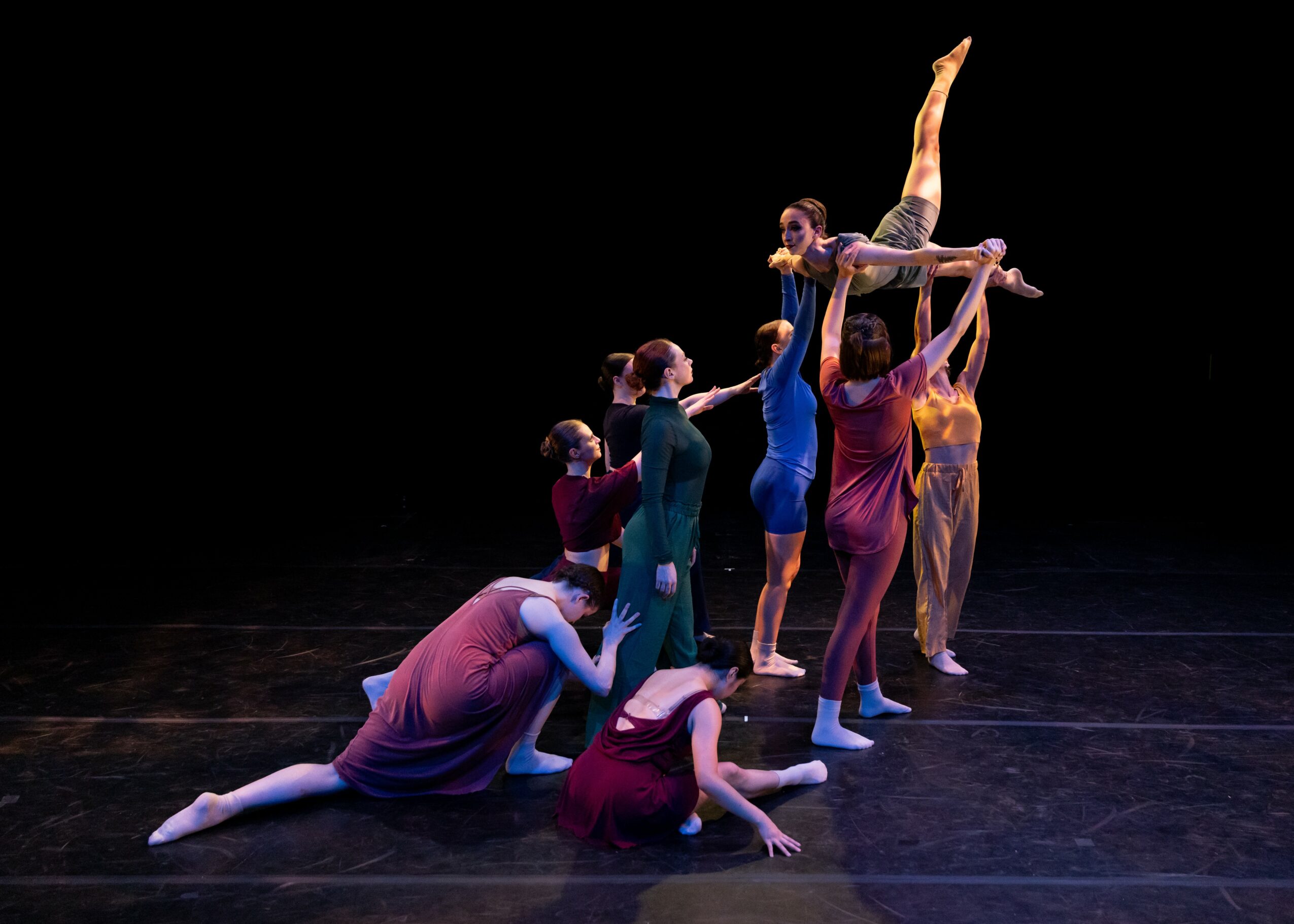 Group of dancers performing a coordinated pose on stage, with one dancer elevated above others against a black background.
