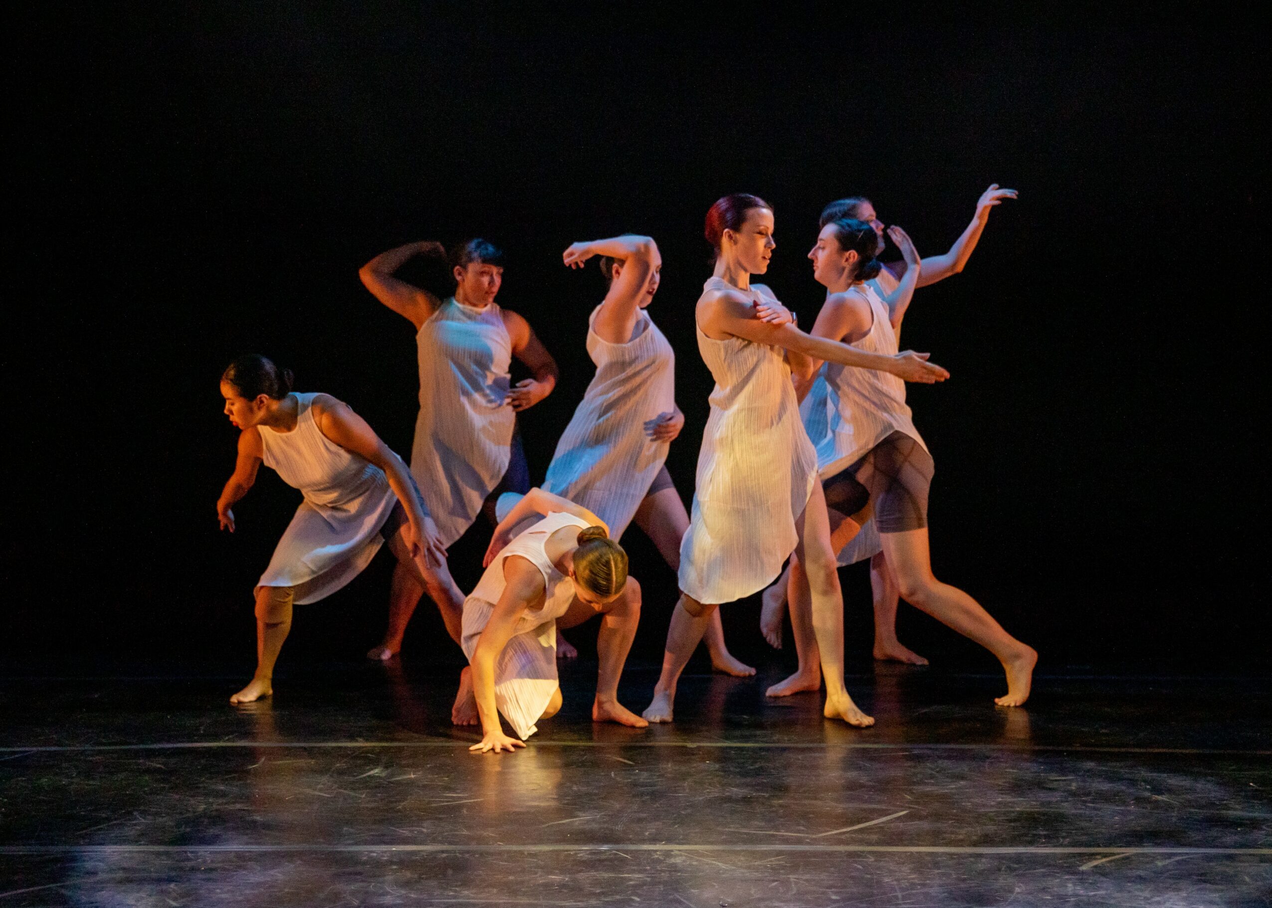 Group of six dancers in white costumes performing on stage with dark background.
