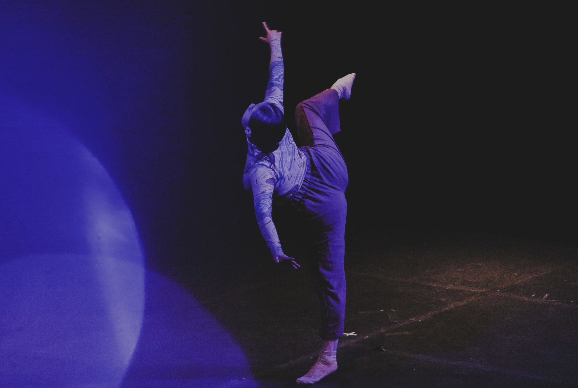 Person performing a dance move on stage with a dark background and blue lighting, one leg raised and arm extended upward.