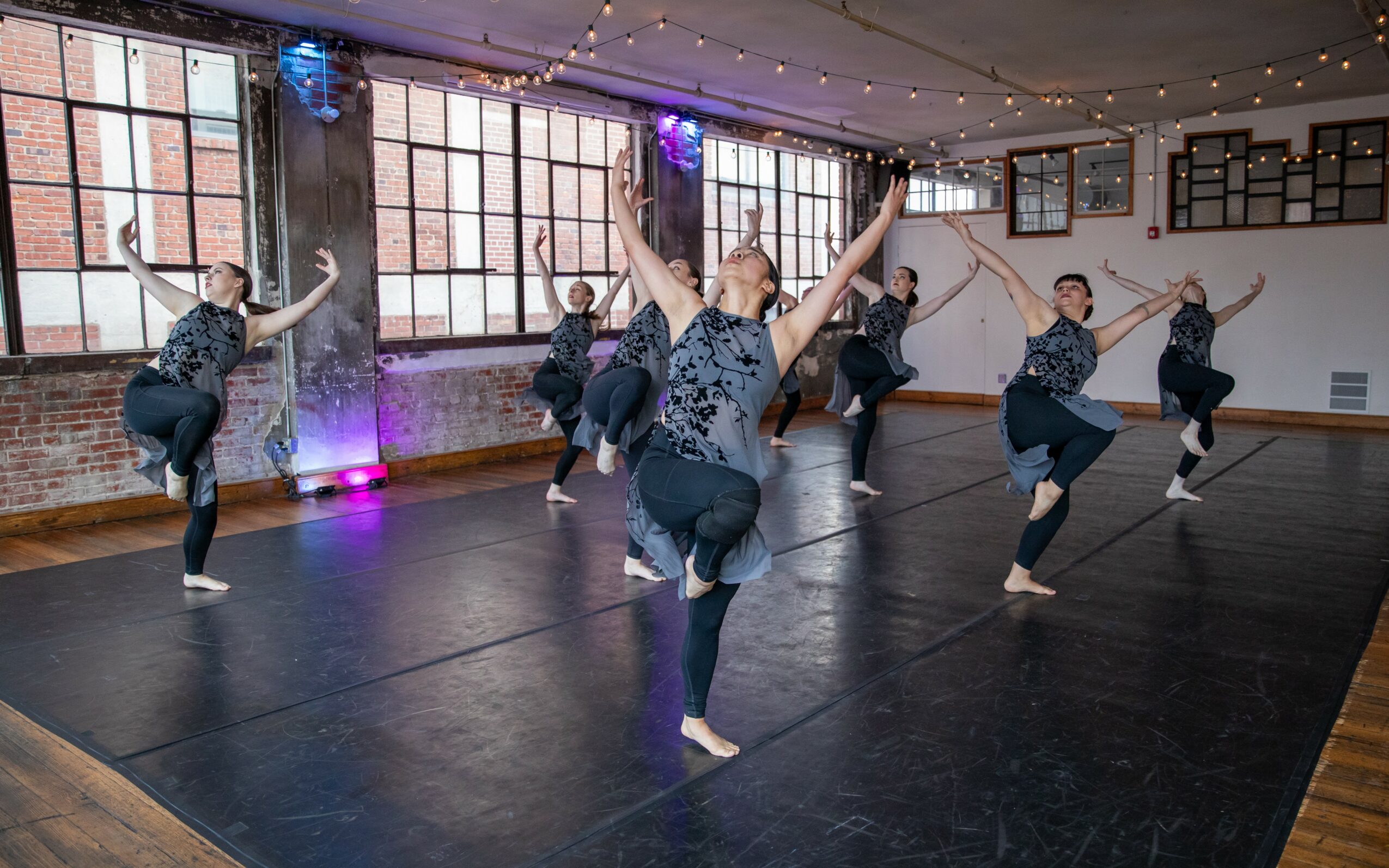 Group of dancers practicing in a studio with large windows and string lights, performing a synchronized move with arms raised.
