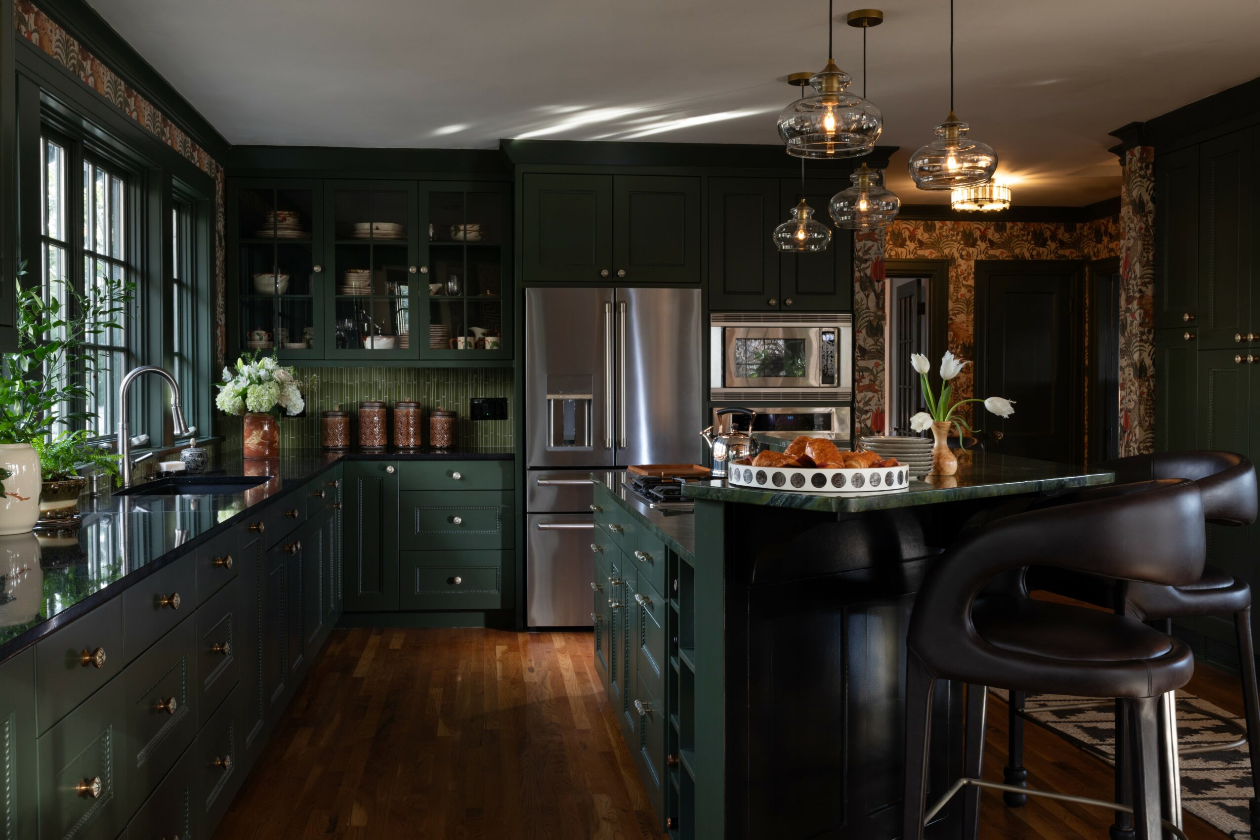 Modern kitchen with dark green cabinets, stainless steel appliances, and a black island with barstool, pendant lights, and wooden flooring.
