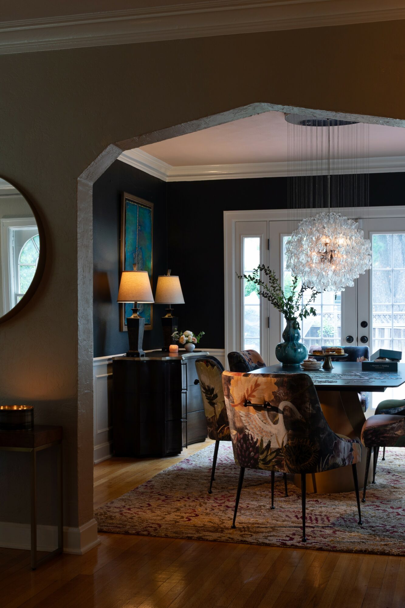 Dining room with chandelier, dark walls, and a large window with glass doors, decorated with flowers and furniture.