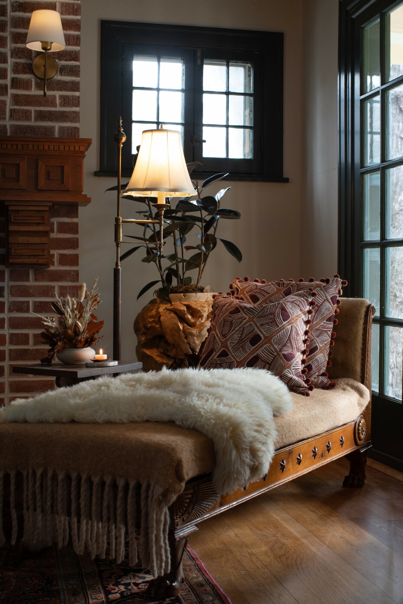 Living room corner with a sofa, pillows, a lamp, a plant, and windows, featuring a brick fireplace and wooden floor.