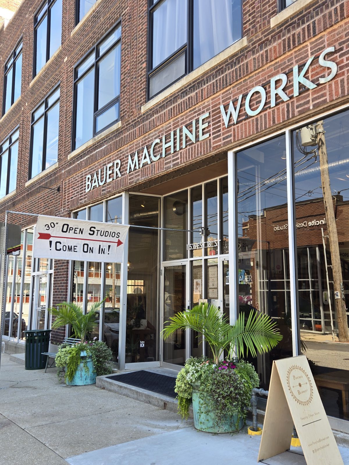 Building with large windows and sign reading 'Bauer Machine Works'. Entrance decorated with potted plants and a sign inviting to open studios.