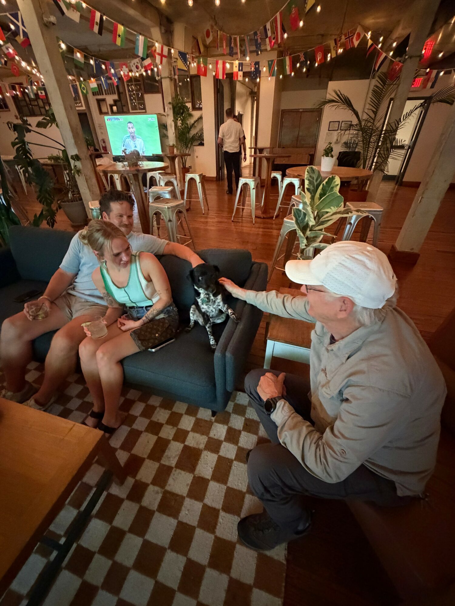 Person in white cap and gray jacket reaching out to a girl in a blue shirt sitting on a black sofa, in a decorated indoor space.