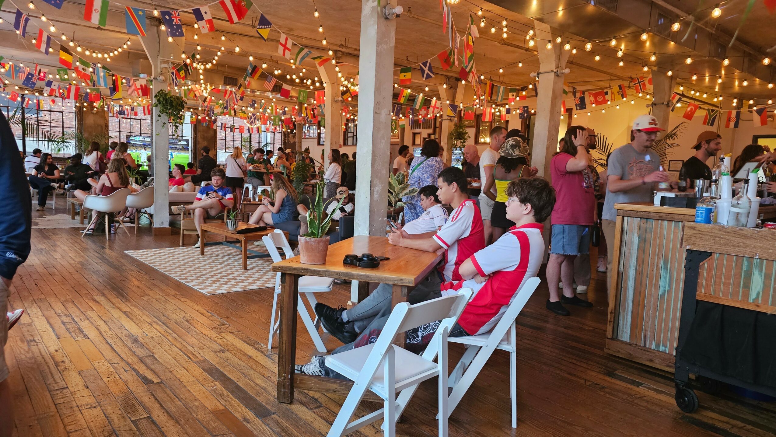 People gather in a decorated indoor space with string lights and colorful banners, sitting at tables and socializing.