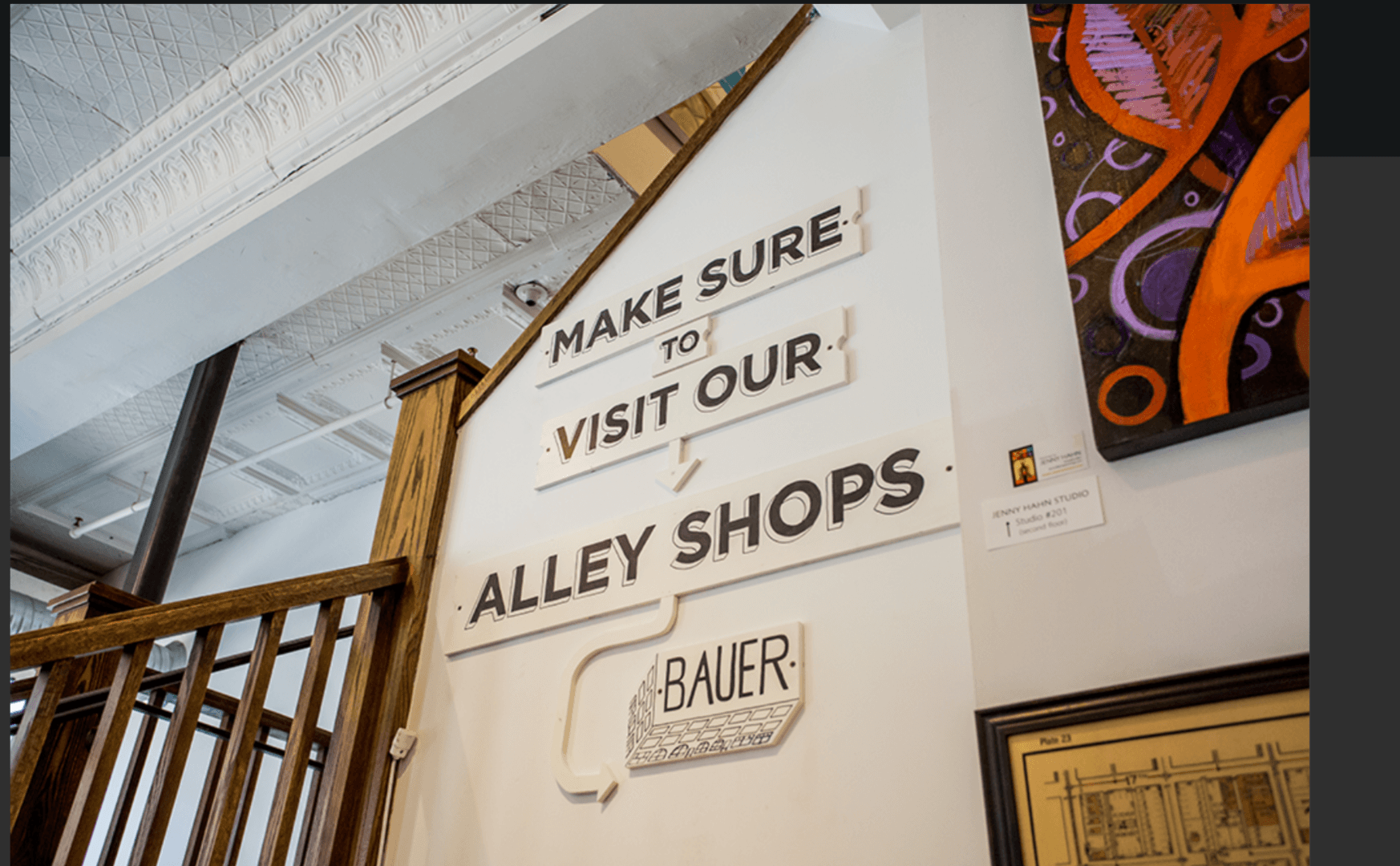 Sign with text: 'Make sure to visit our alley shops' in a gallery or shop setting.