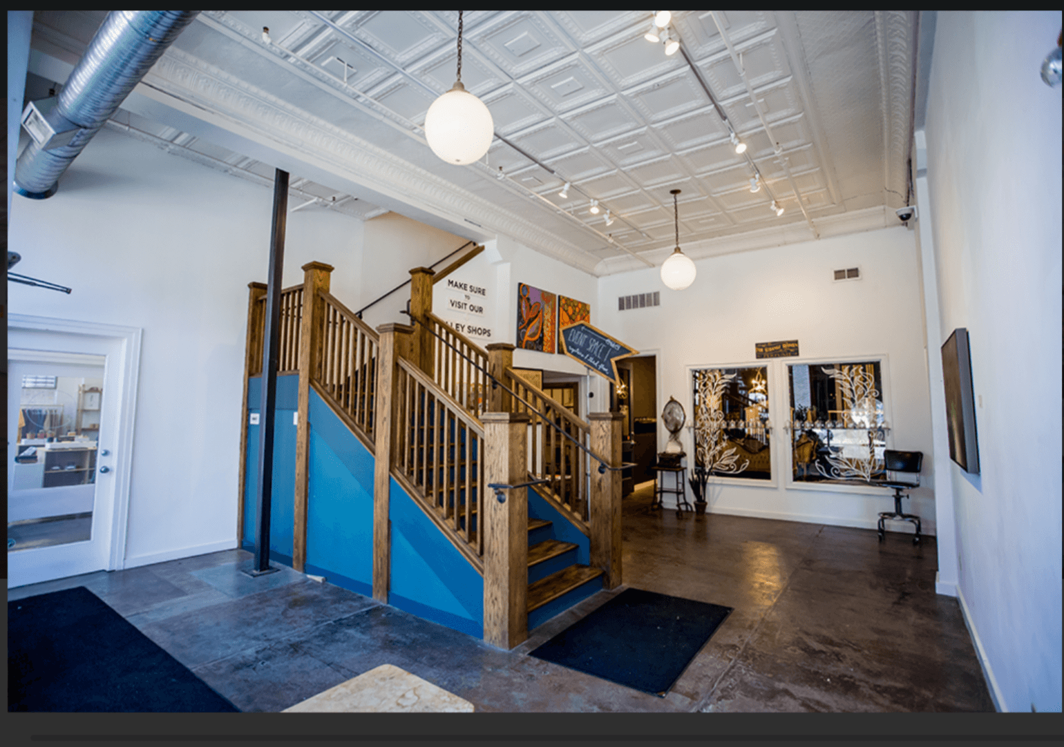 Interior space with wooden staircase, artwork on walls, and large windows, high ceiling with hanging lights, polished floor.