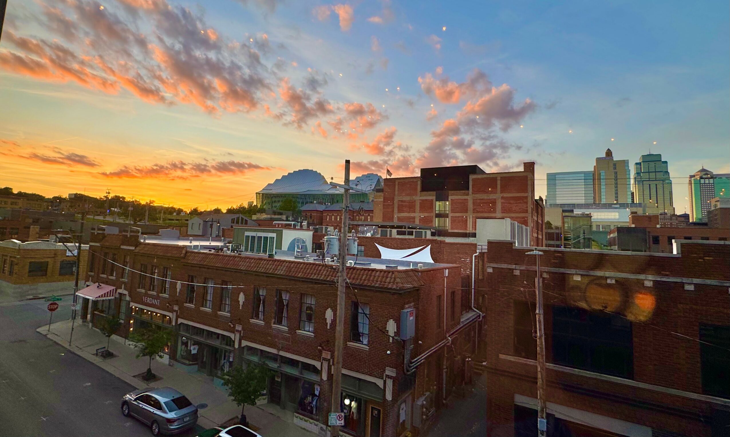 Cityscape with brick buildings and a sunset sky with clouds, trees, and cars on the street.