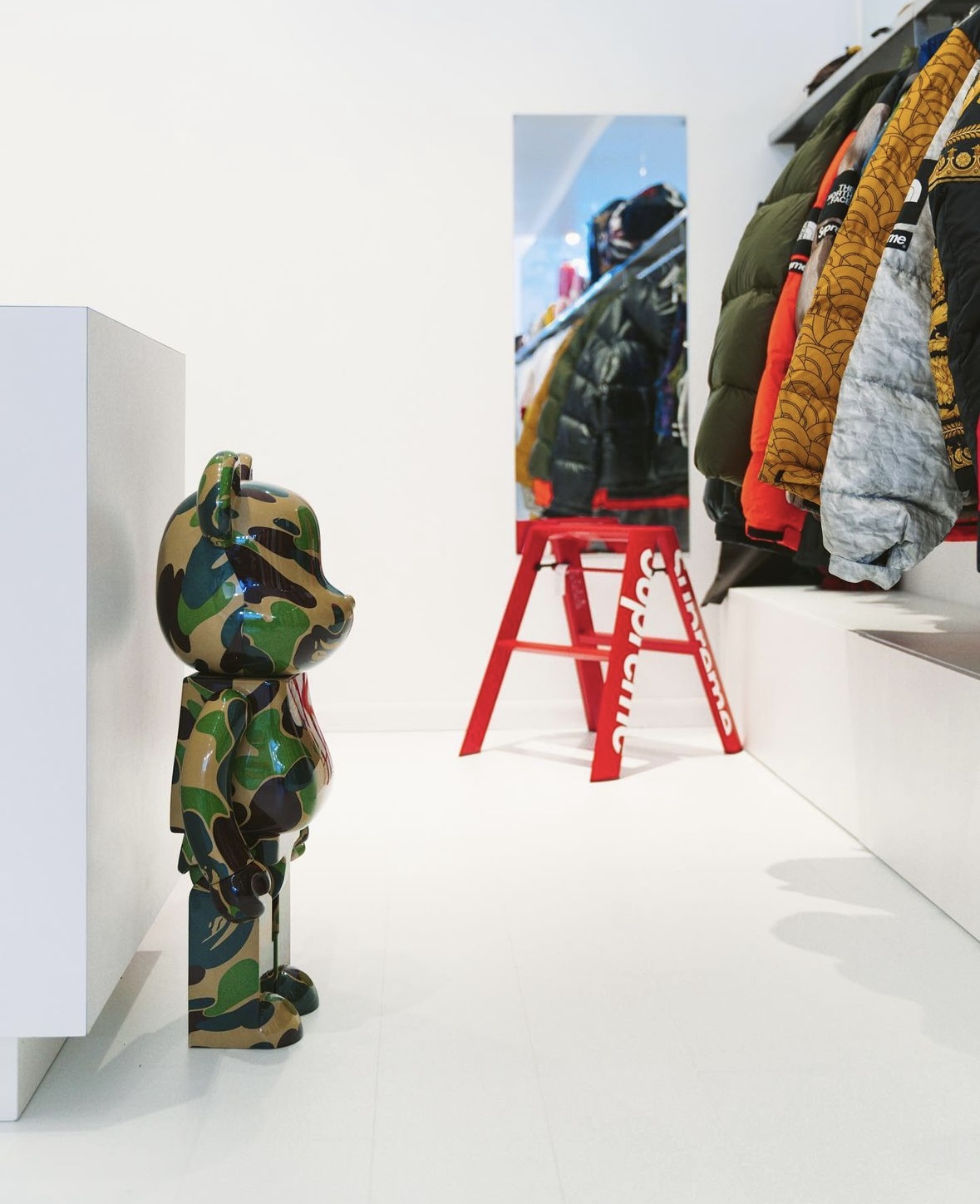 Child-sized camouflage coat hanging on a hook, with a mirror and coat rack in the background, in a clothing store.