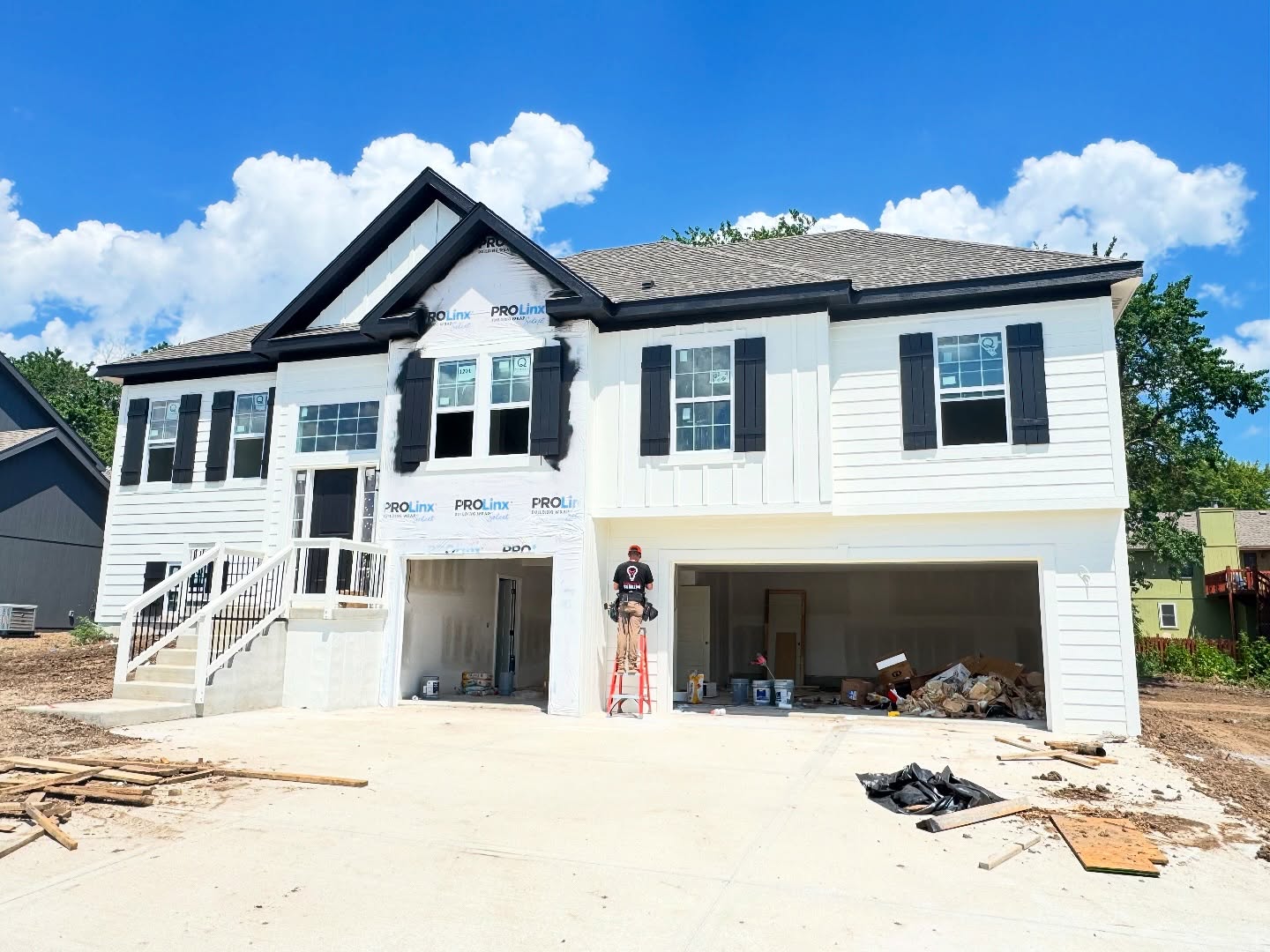 New two-story house under construction with white siding, black shutters, and a garage, on a sunny day with blue sky.