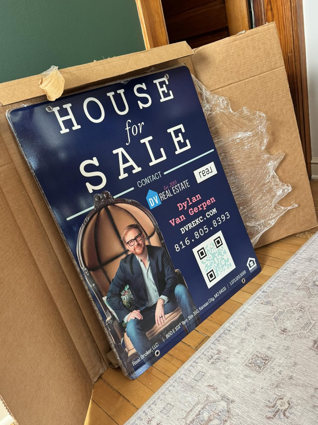 House for sale sign with a woman sitting on a chair, contact details, and a QR code, leaning against cardboard boxes.