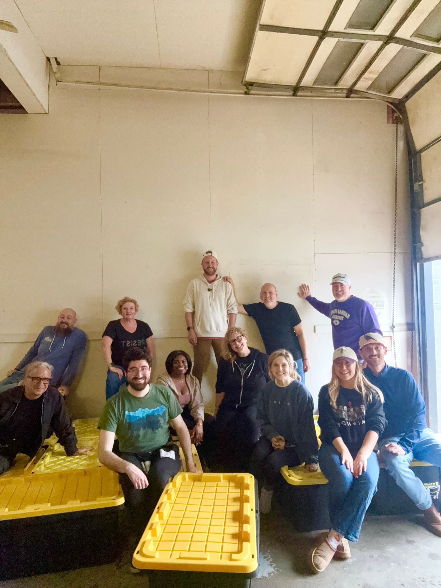 Group of ten people sitting and standing in a warehouse, some smiling, with yellow crates in front, wall and garage door in background.