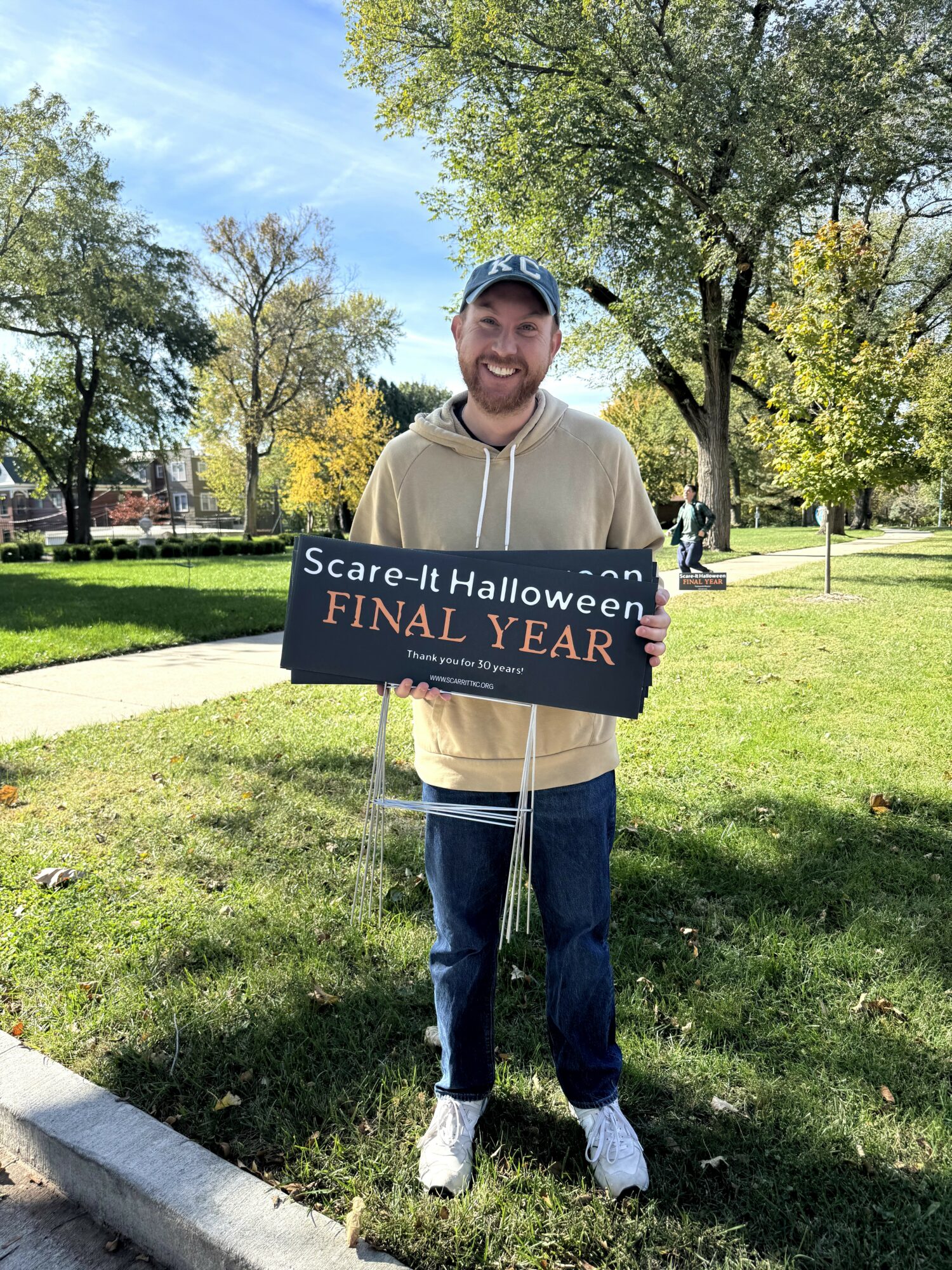 Smiling person holding a sign in a park with trees and grass, sunny day, wearing a hoodie, jeans, and sneakers.