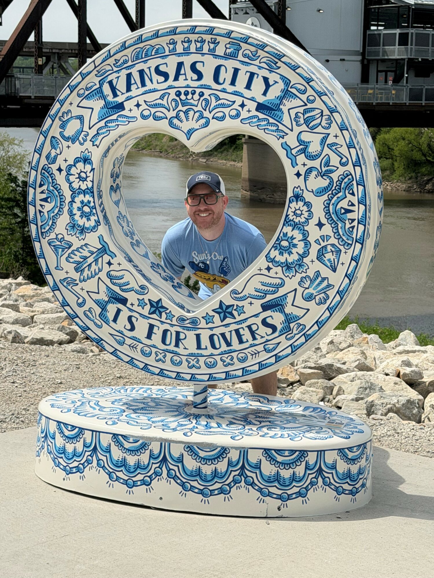 Person smiling inside a heart-shaped Kansas City sign with blue floral patterns, outdoors near water and rocks.