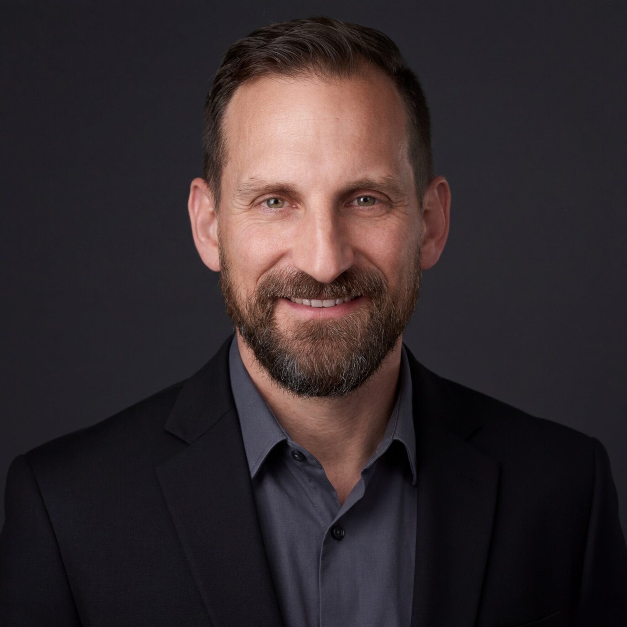Portrait of a smiling man with short dark hair and a beard, wearing a dark suit and shirt, against a dark background.