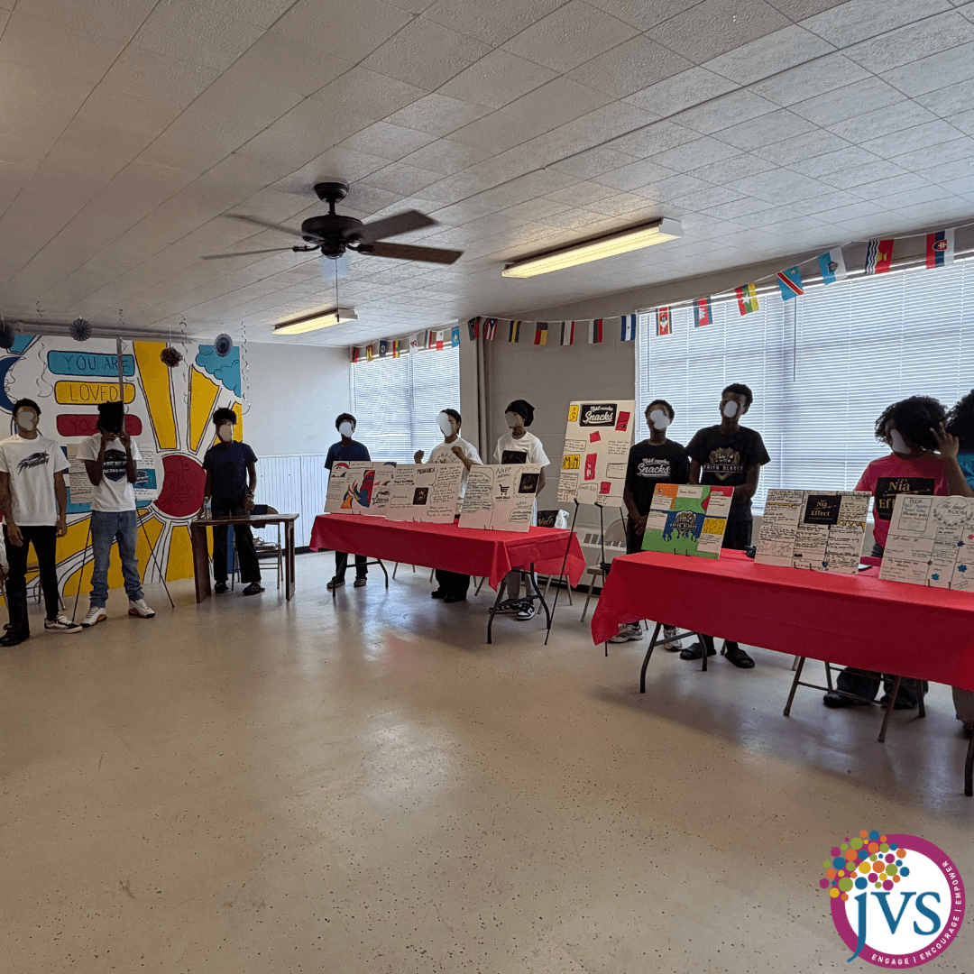 People standing behind tables with displays in a decorated room, some wearing masks, colorful banners hanging, and a mural on the wall.