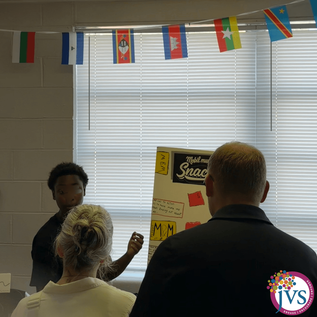 Two children and two adults in a classroom with international flags and a window with blinds.