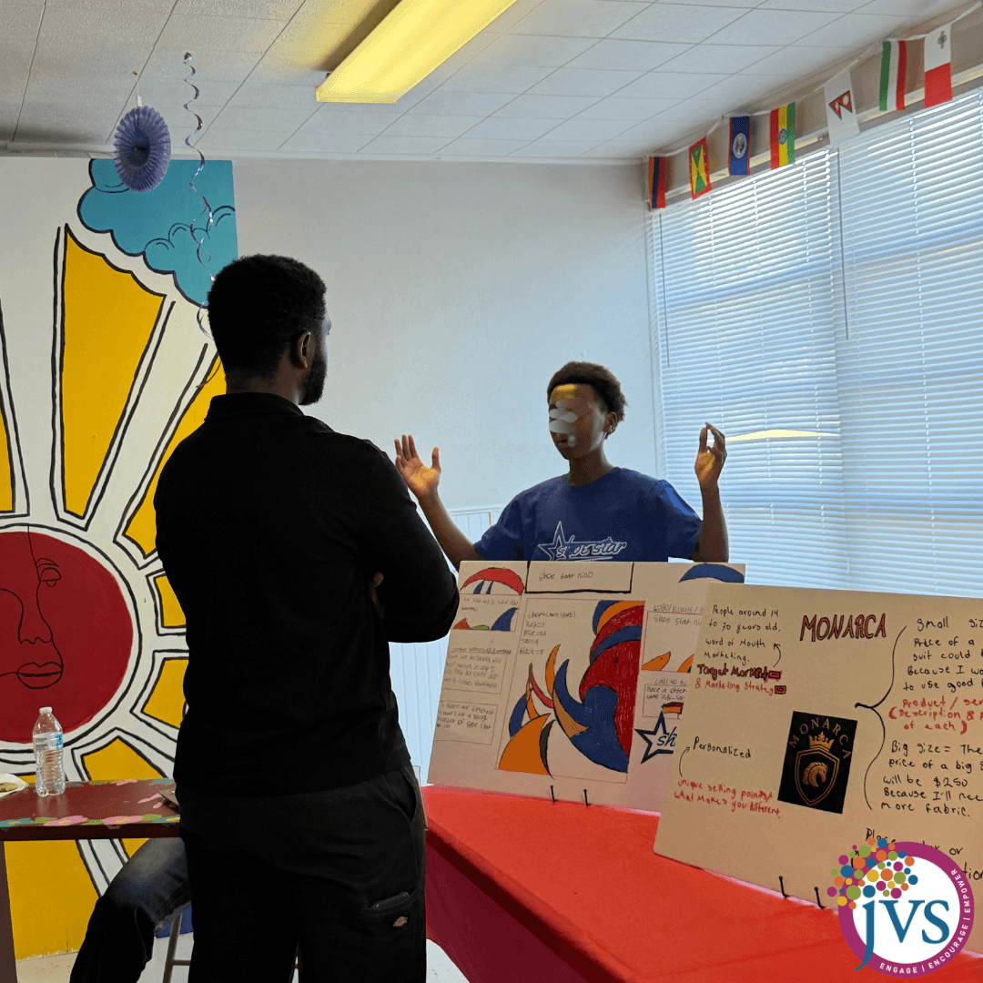 Two people stand in a room with display boards, flags, and a colorful wall decoration, engaging in conversation.