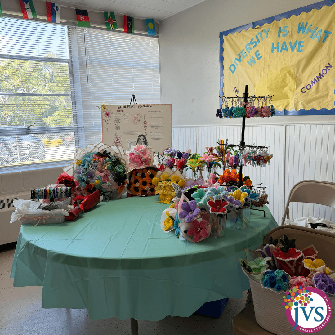 Table with colorful handmade flowers and crafts in a room with window and educational posters, decorated for an event.