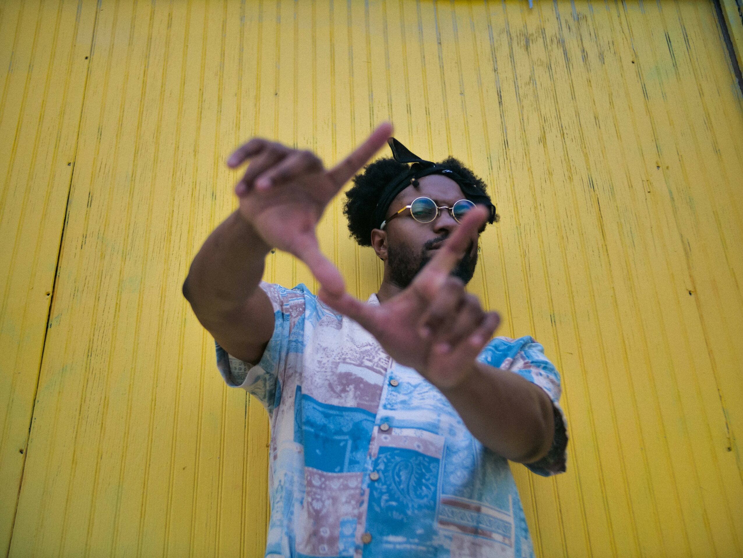 Person with glasses and dreadlocks making a hand gesture against a yellow background.