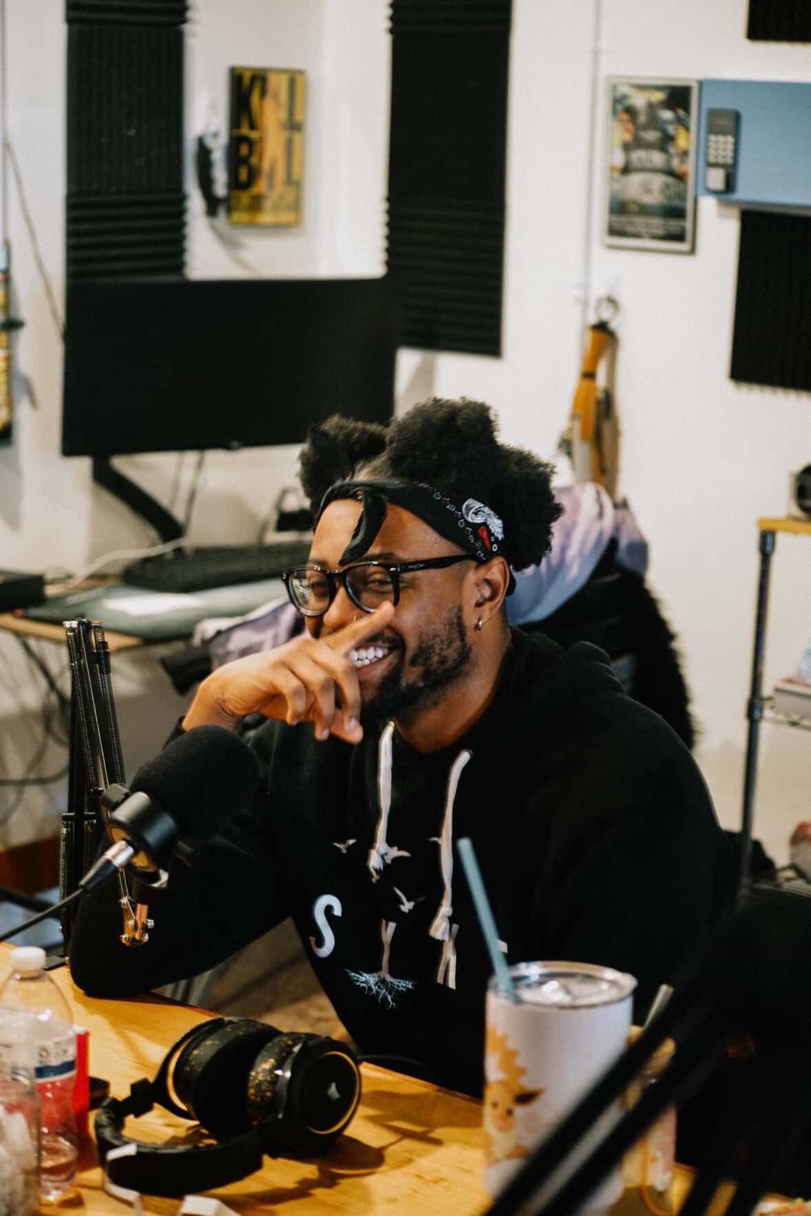 Man with glasses and a beard smiling, sitting at a table in a recording studio, with headphones and microphone, surrounded by equipment.