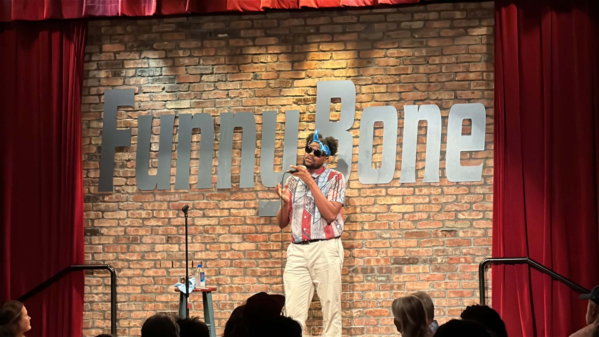 Person performing stand-up comedy on stage with brick wall background and large 'Funny Bone' sign, audience visible.