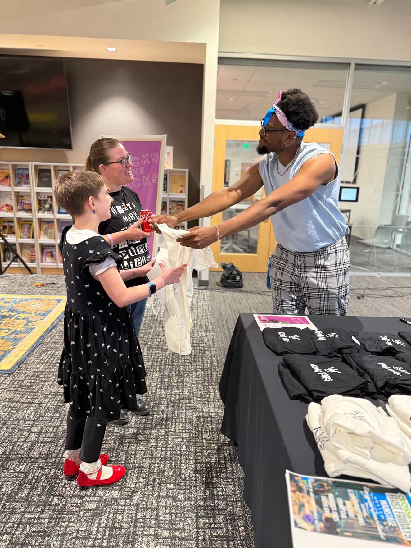 A person with curly hair and glasses hands an item to a young girl in a black dress with white polka dots, in an indoor setting.