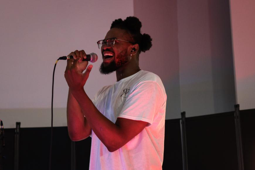 Person singing into a microphone, smiling, with a dark background and pinkish lighting.
