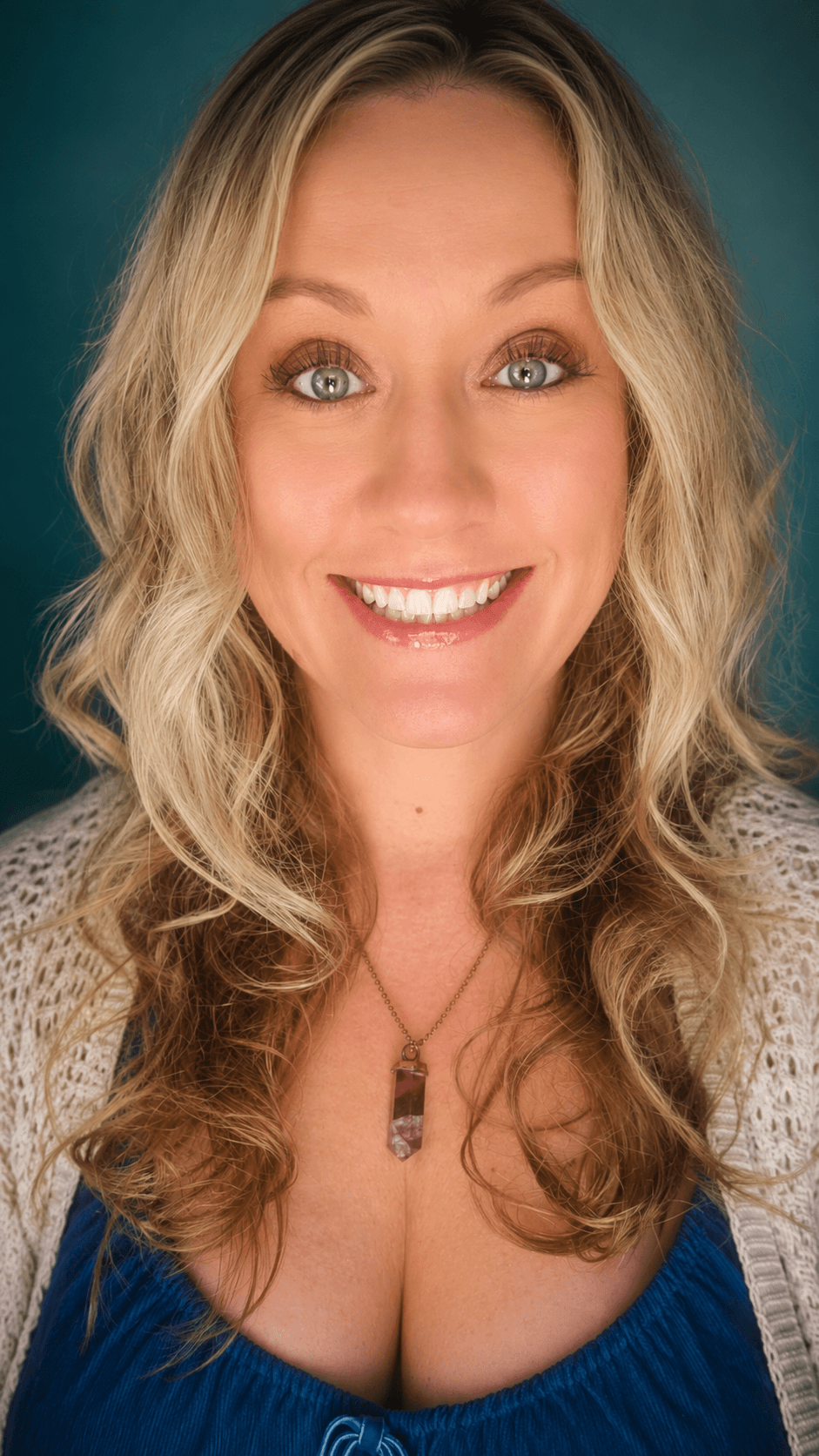 Smiling woman with wavy blonde hair wearing a blue top and necklace, against a teal background.