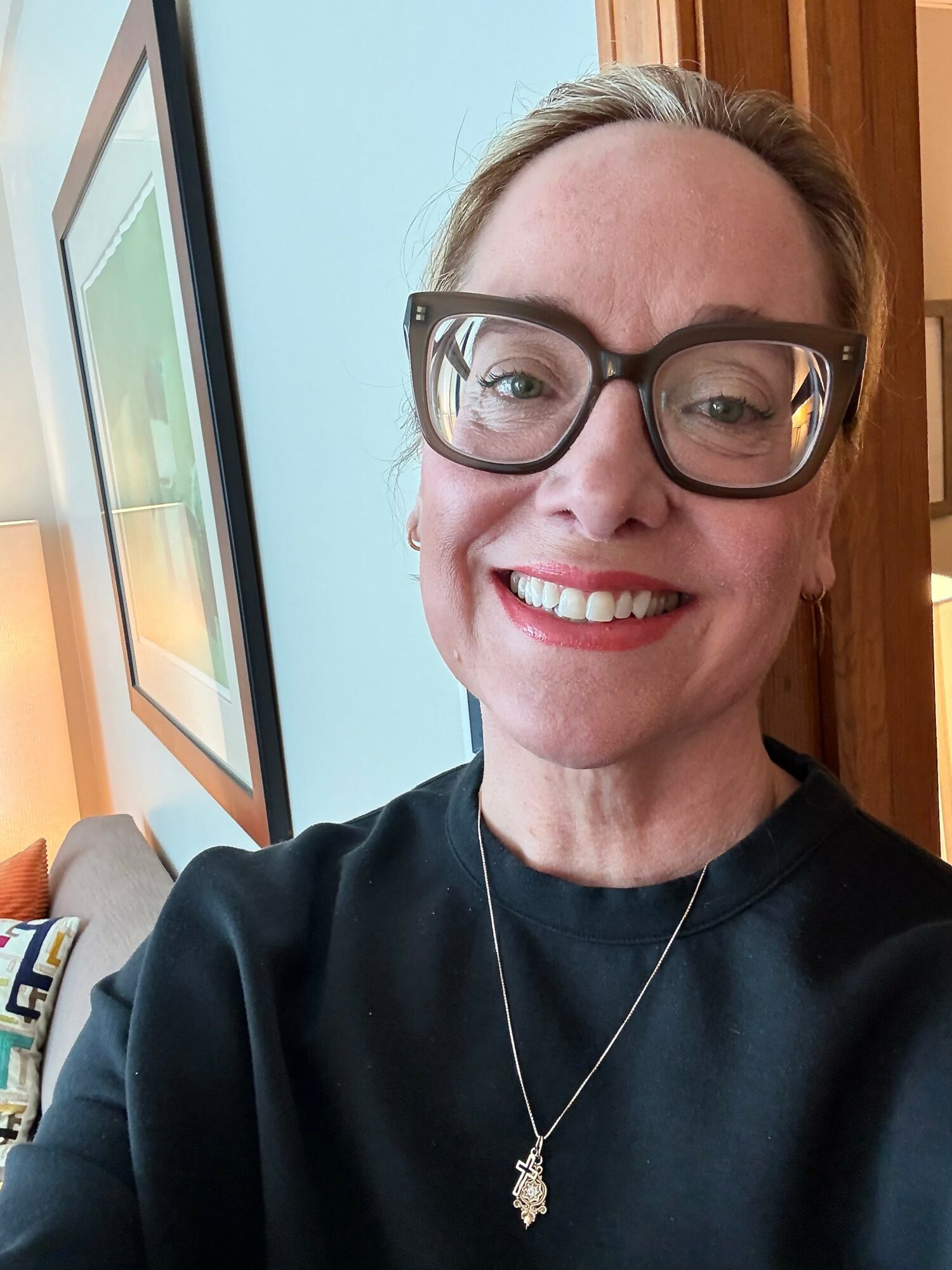 Woman with glasses smiling, wearing a black top and necklace, in a room with a framed picture on the wall.