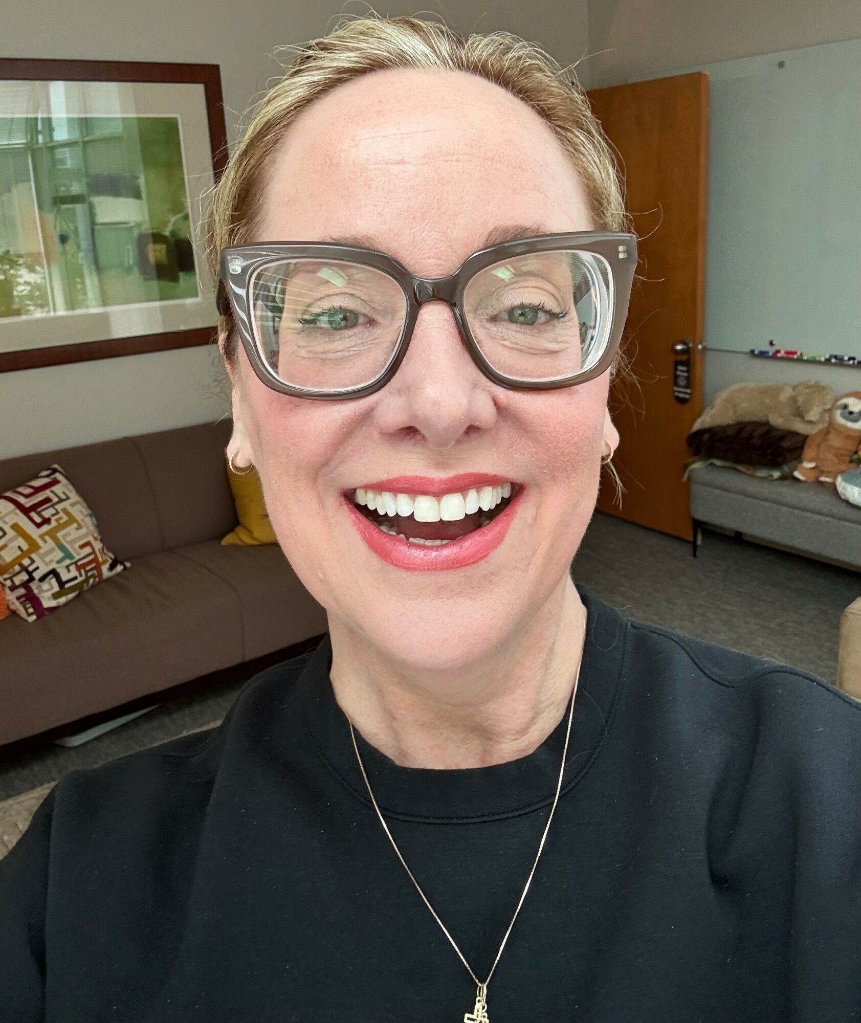 Smiling woman with glasses and a necklace, indoors with a couch and framed picture in the background.