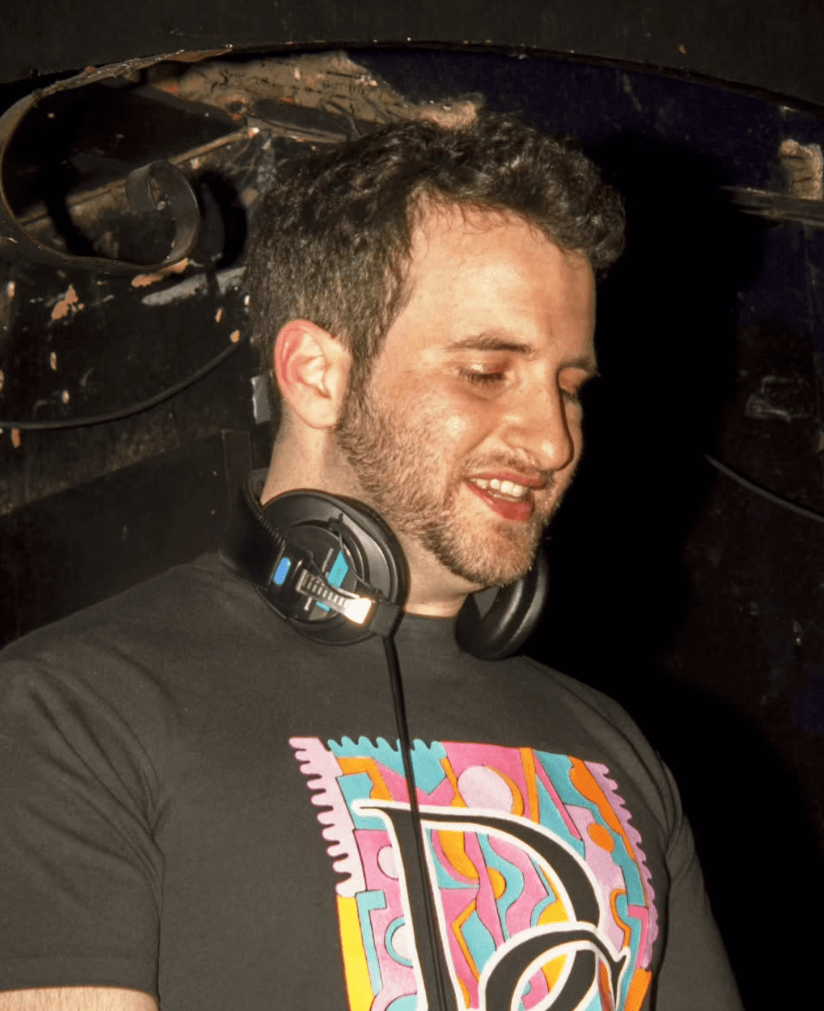 Young man with headphones smiling, wearing a black T-shirt with colorful design, dark background.