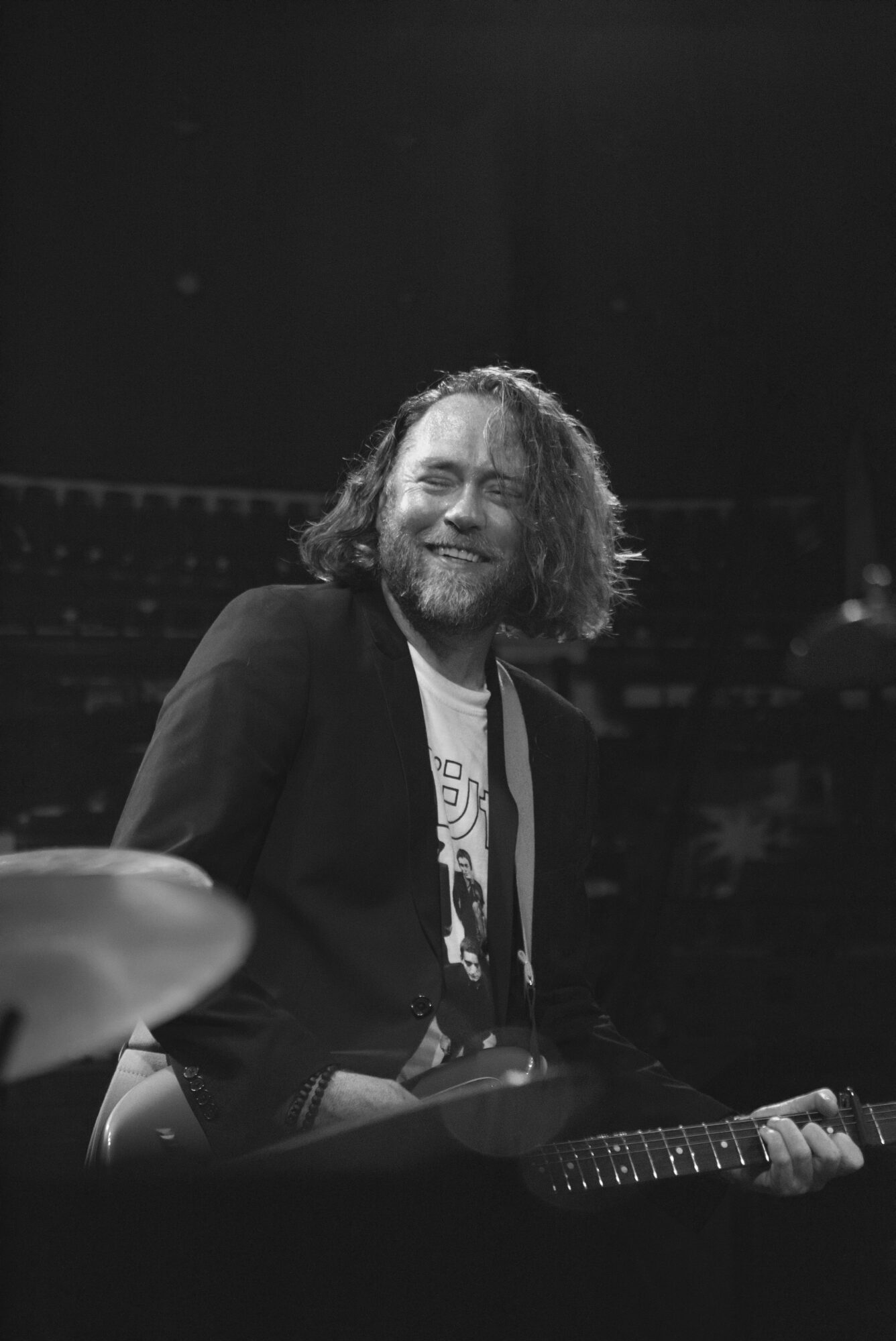 Man with curly hair and beard smiling, holding a guitar, in a dark setting.