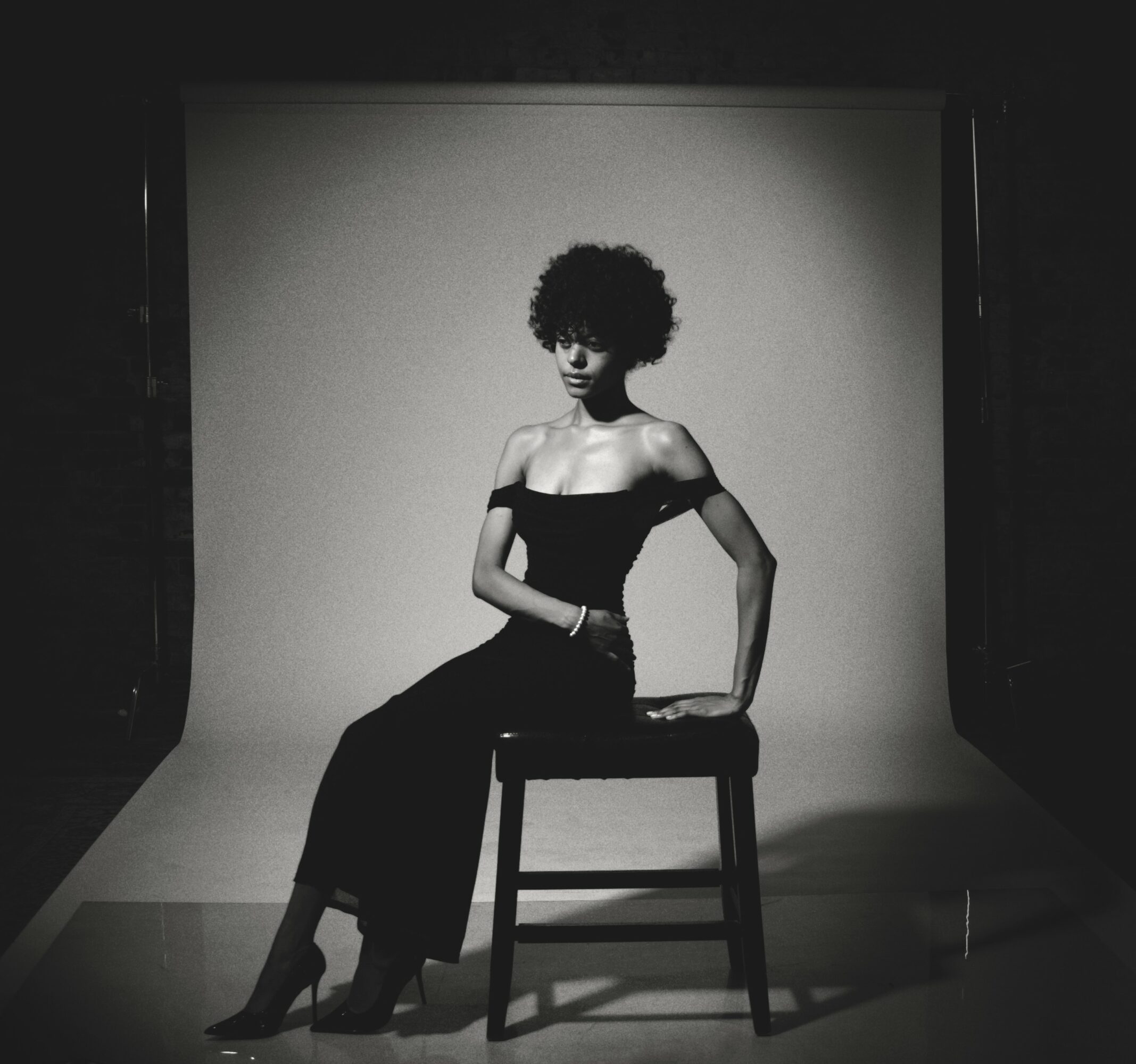 Woman with curly hair sitting on a chair in a photography studio, wearing a strapless dress, in black and white.