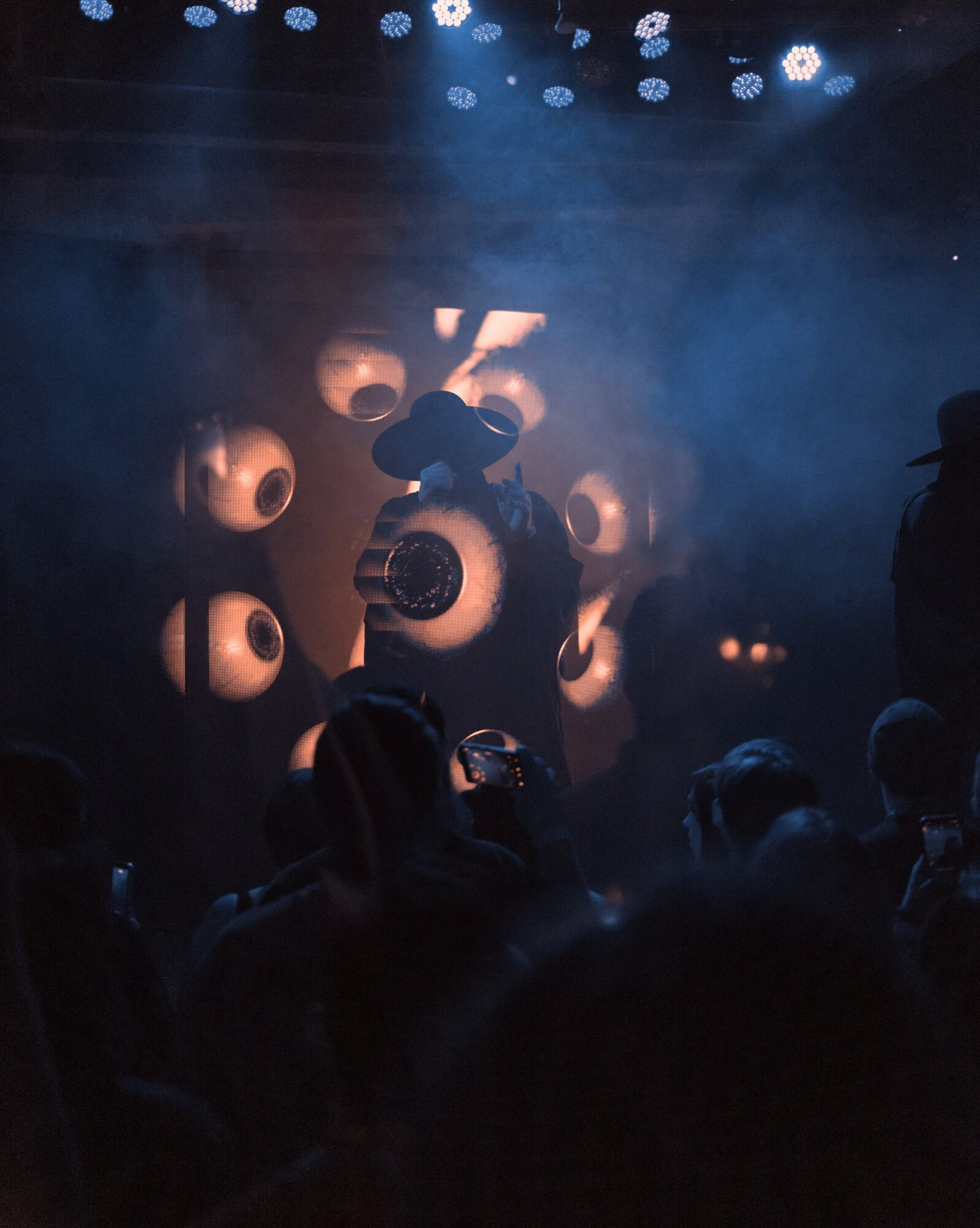 Stage with fog, large circular lights, and a performer with a hat, audience silhouettes visible, dim lighting.
