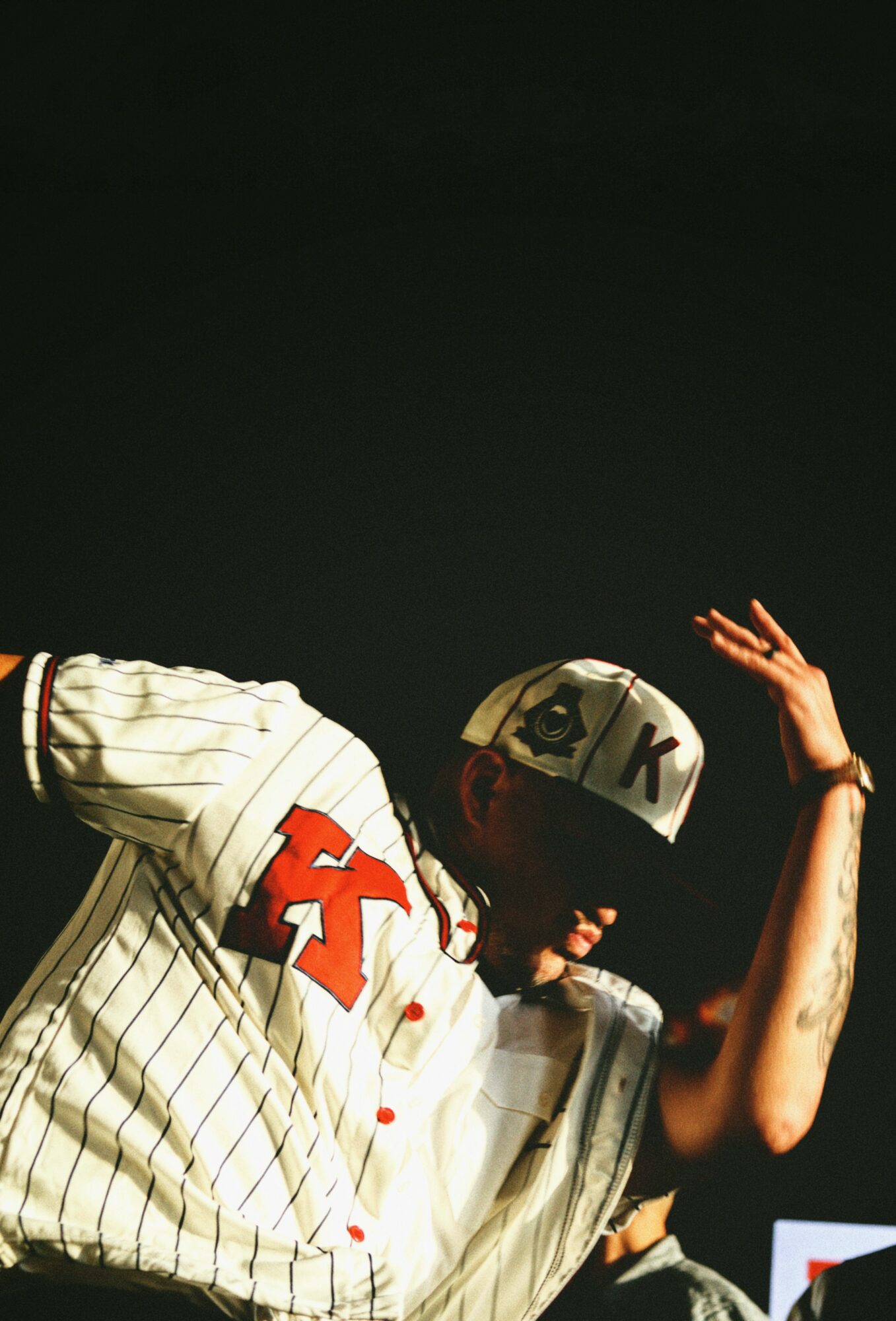 Person wearing a baseball jersey and cap, raising their right hand, against a dark background.