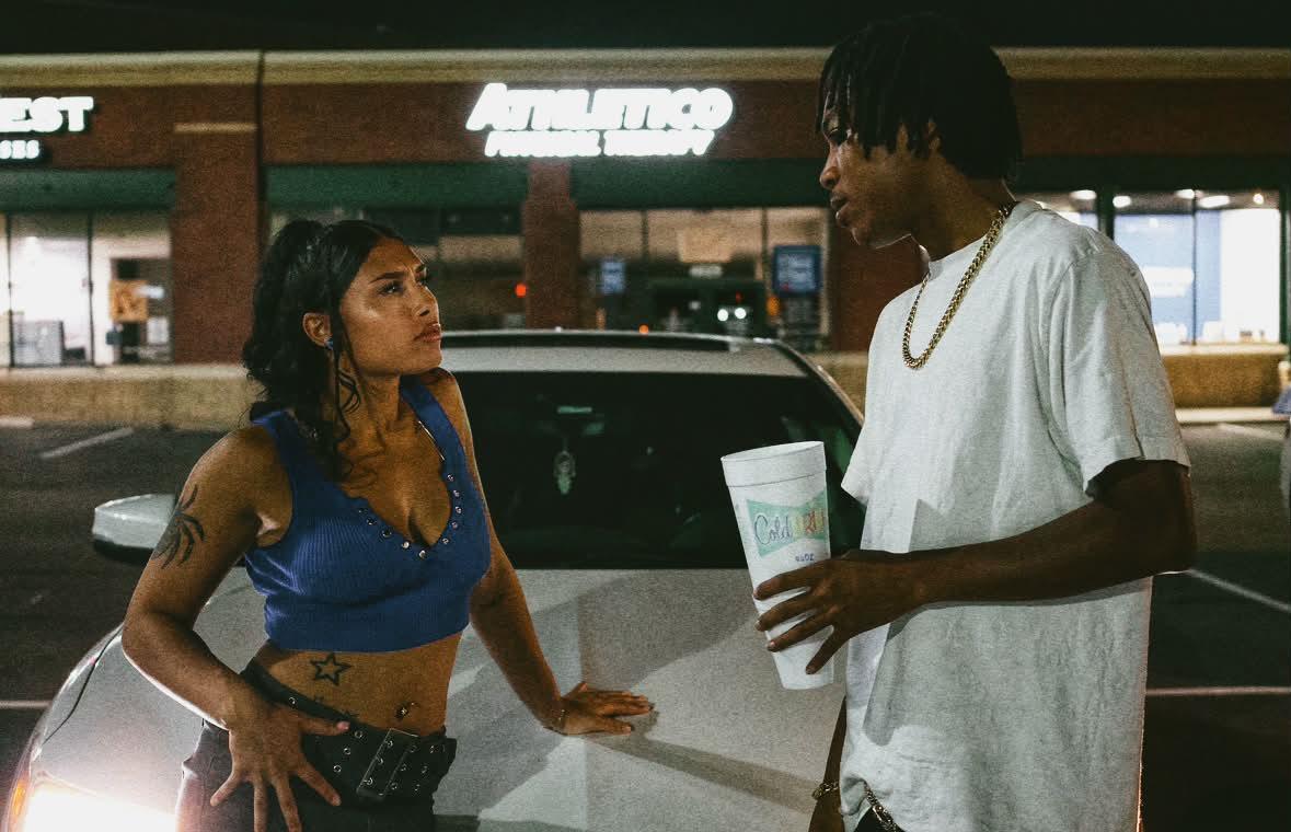 A woman and a man are talking outside a store at night, with a car behind them and store signs visible.