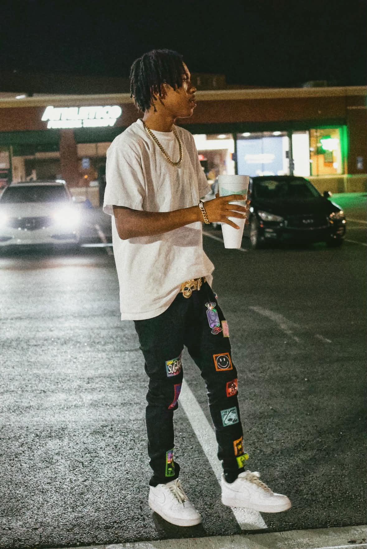 Young man with dreadlocks holding a drink in a parking lot at night, wearing a white t-shirt, black pants, and white sneakers.