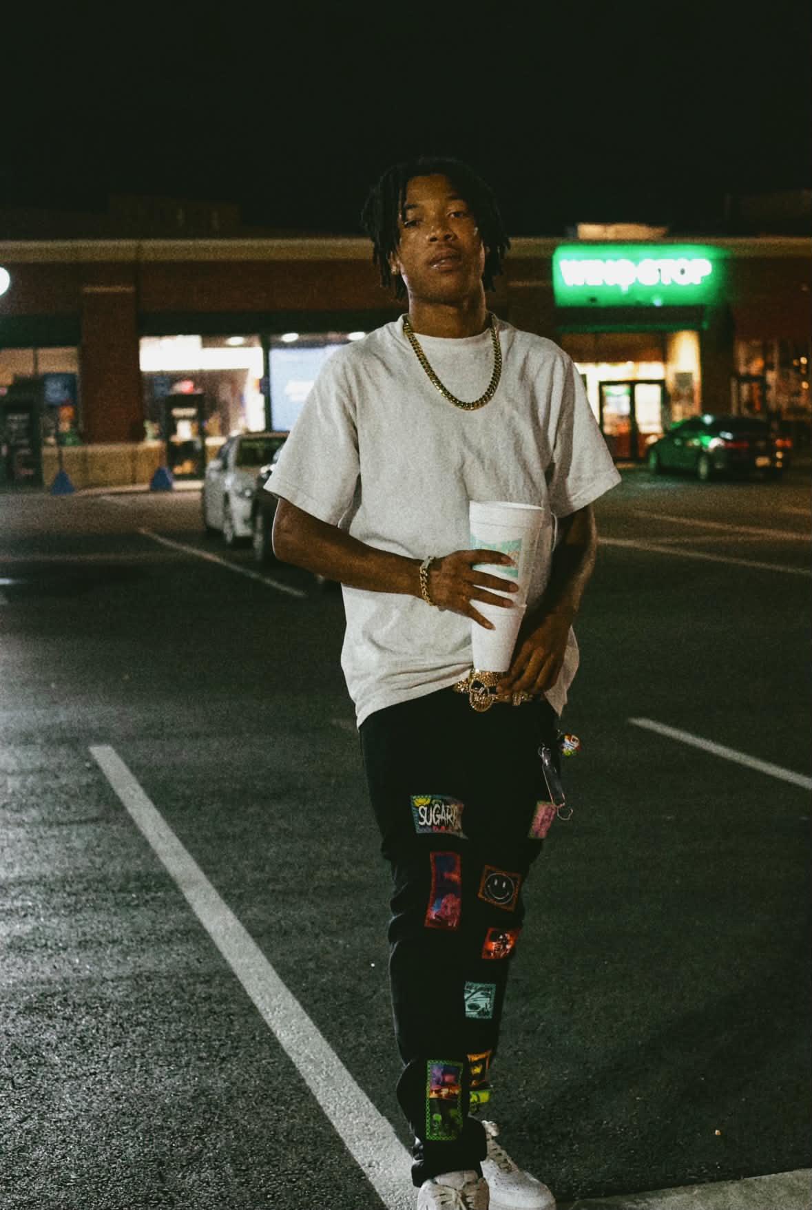 Person standing in a parking lot at night holding a drink, wearing a white t-shirt and patterned pants.