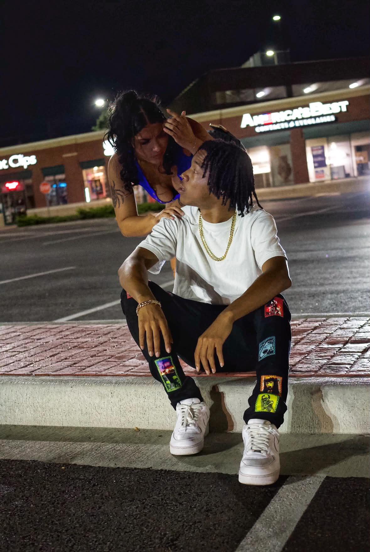 Two people sitting on a curb at night, one leaning in to the other, with a shopping center in the background.