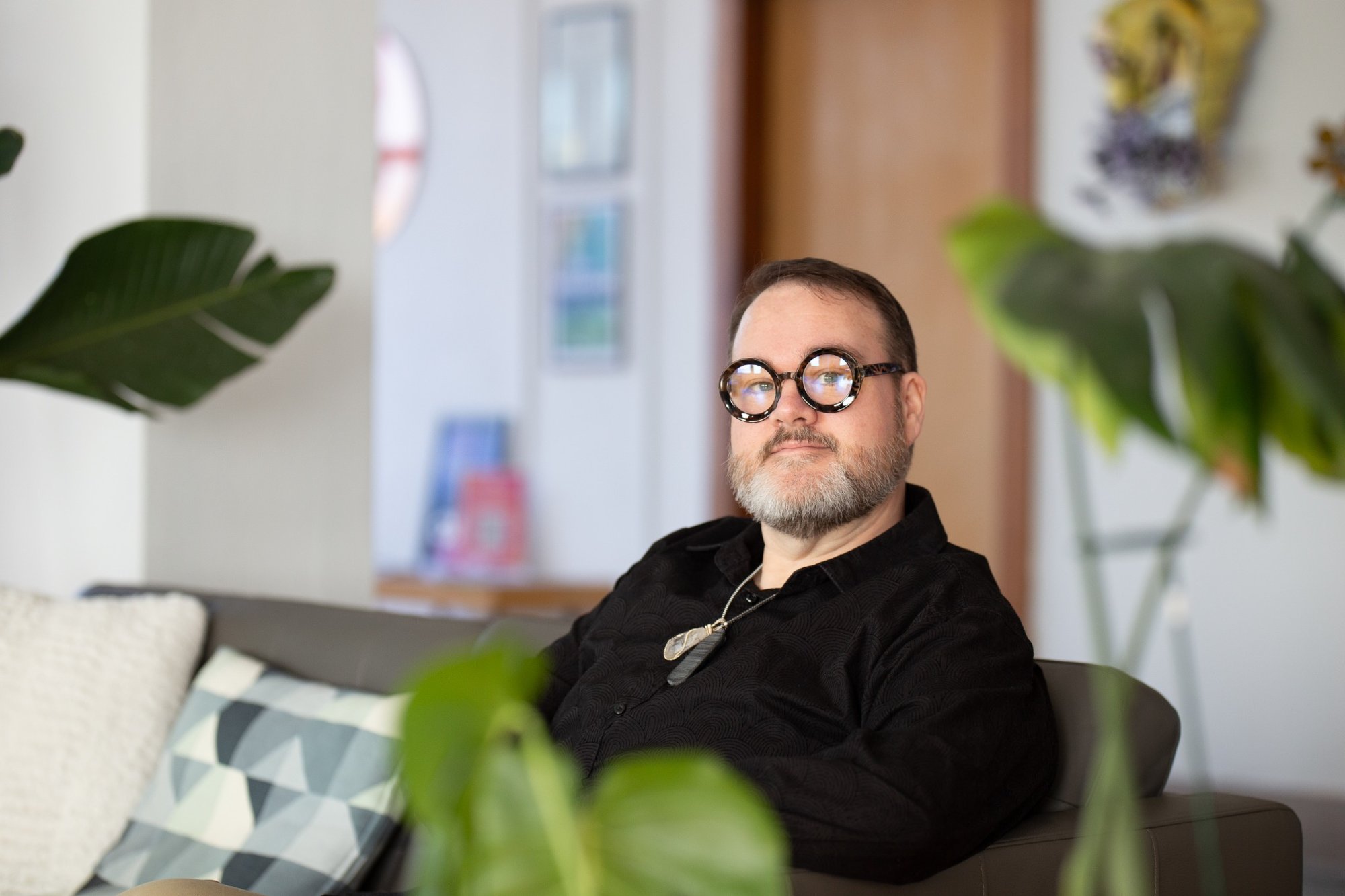 Man with glasses and beard sitting on a sofa in a room with plants and colorful decor