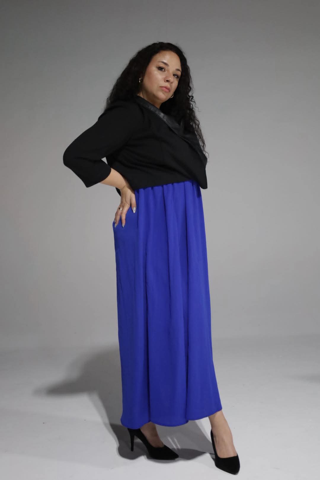 Woman with long dark hair wearing a black top and blue wide-leg pants, standing with hand on hip, facing camera.