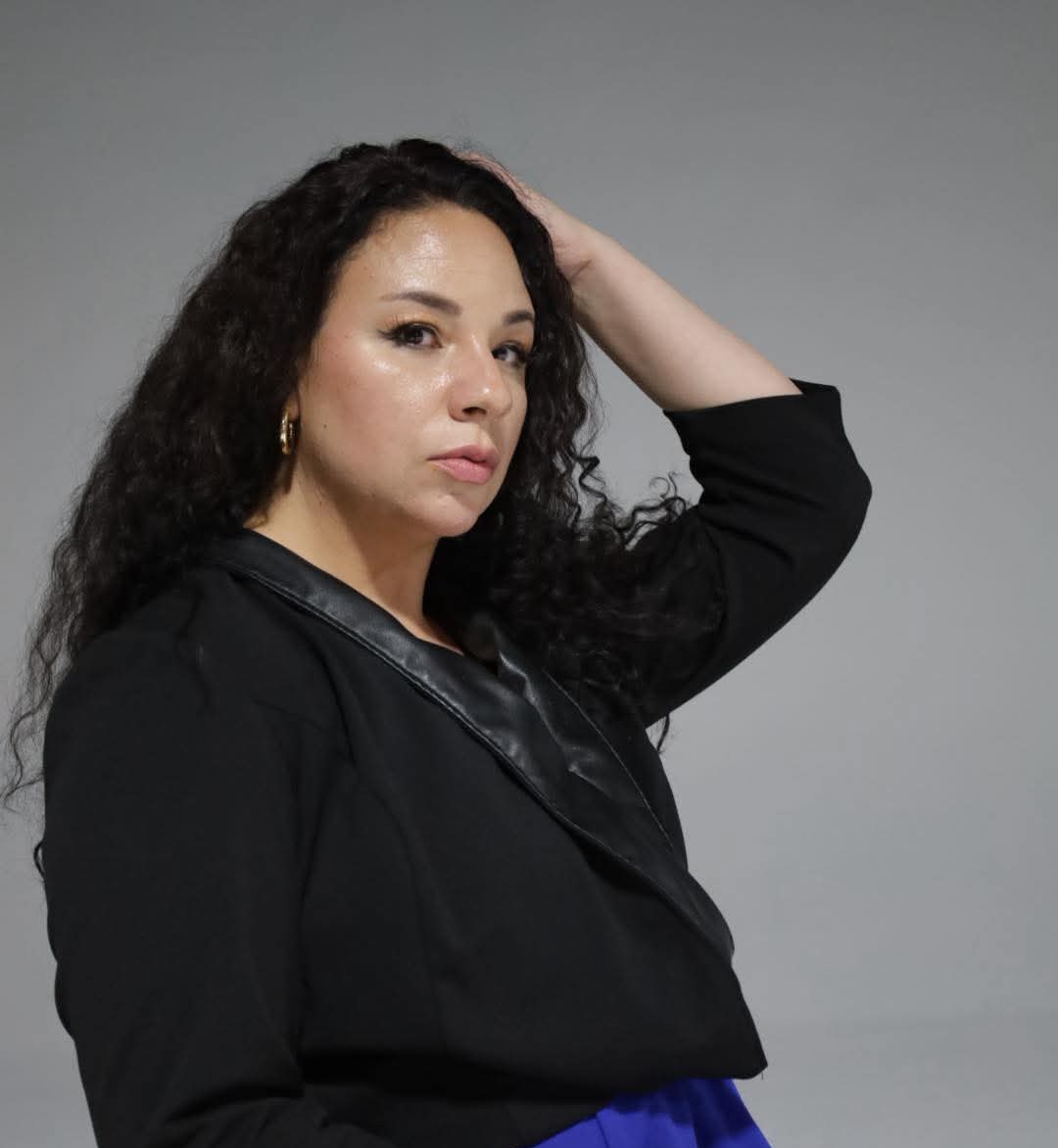 Woman with curly hair wearing a black jacket, posing with her hand on her head, against a plain gray background.