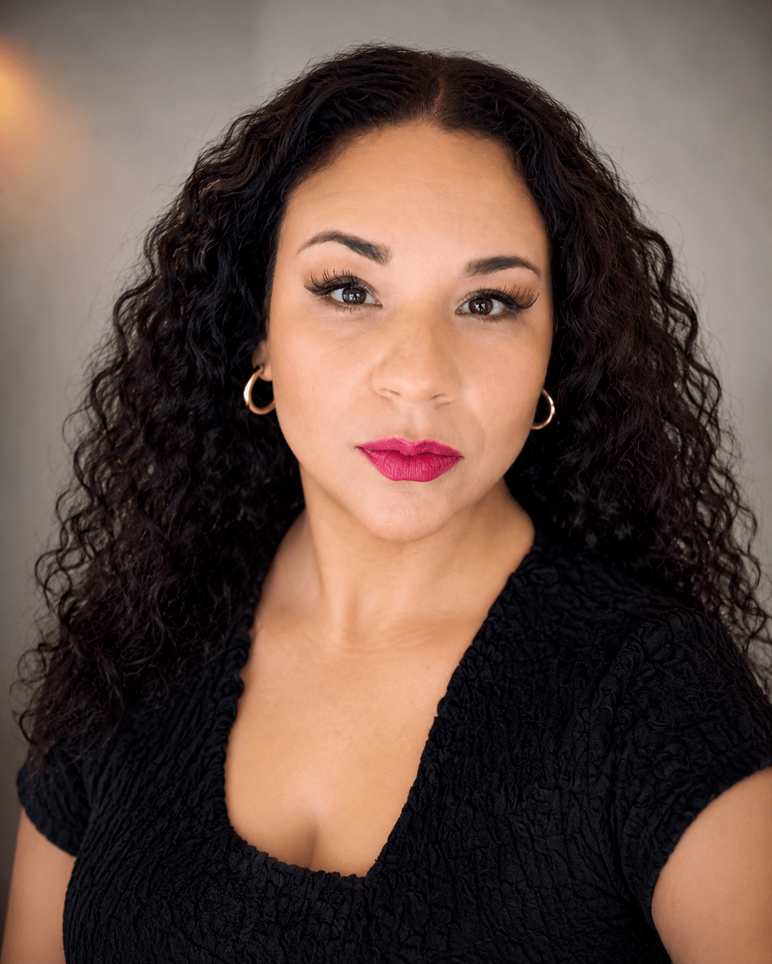 Portrait of a woman with curly hair, wearing a black top and gold hoop earrings, looking directly at the camera.
