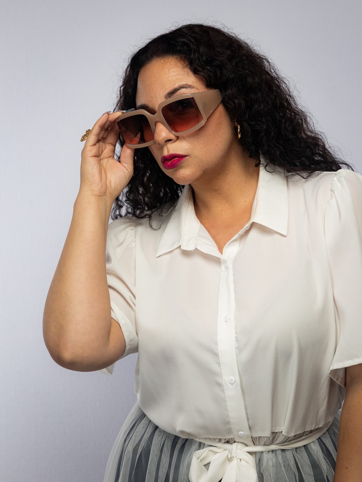 Woman with curly hair wearing sunglasses and a white blouse, adjusting her glasses, against a plain background.