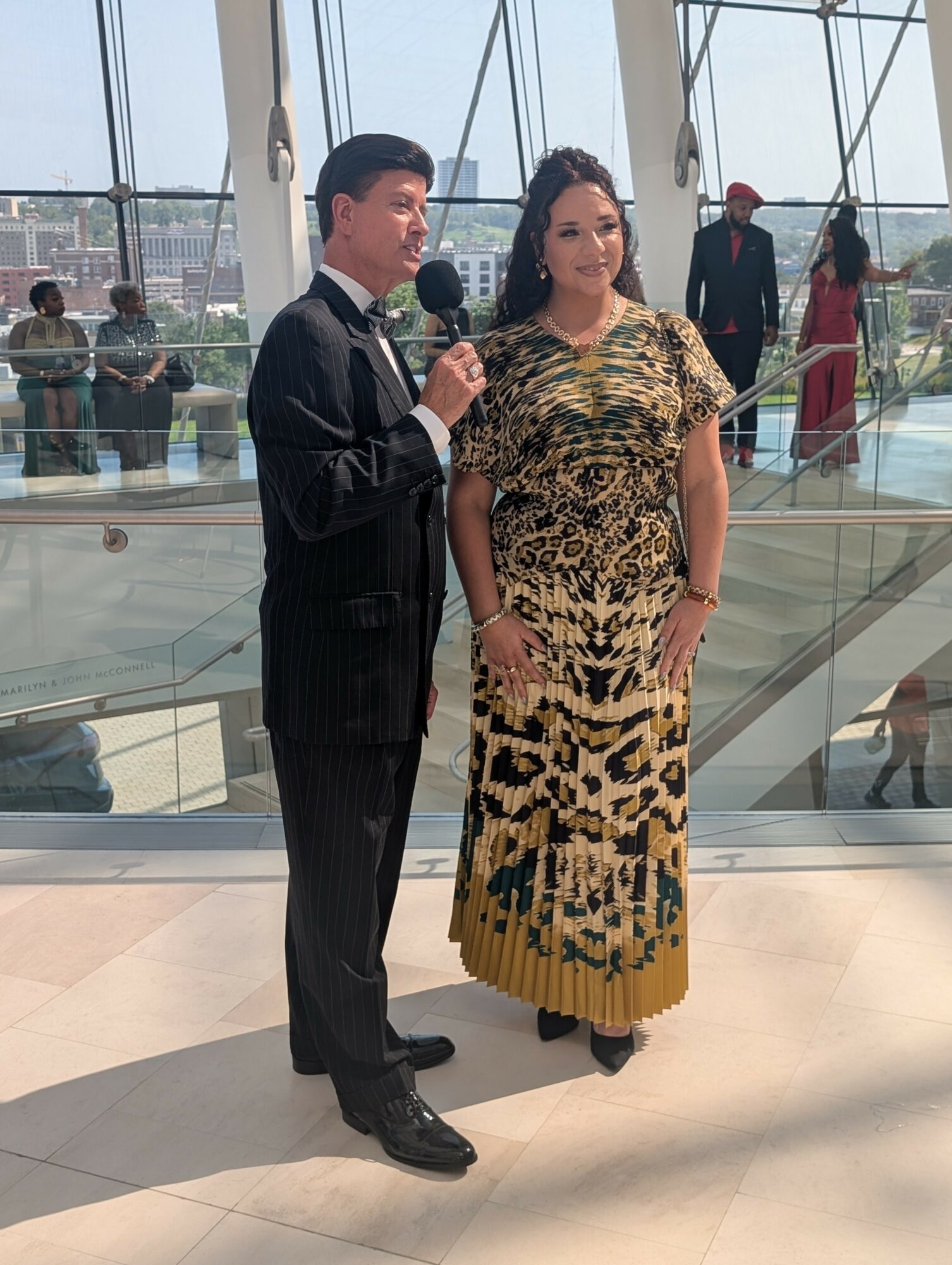 Man and woman standing indoors, woman wearing a patterned dress, man holding a microphone, glass railing behind them.