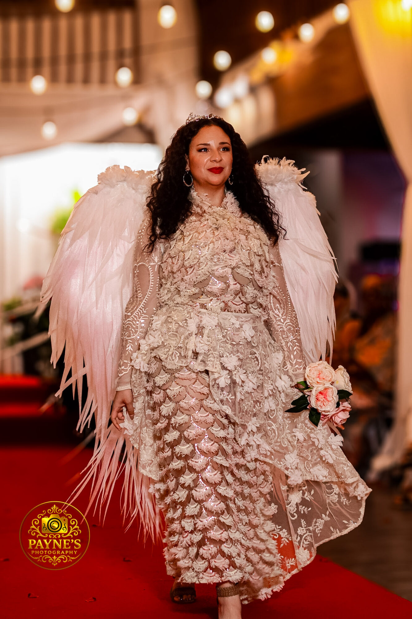 Woman in ornate dress with large pink feathered wings holding a bouquet walks on red carpet at event, background with lights.